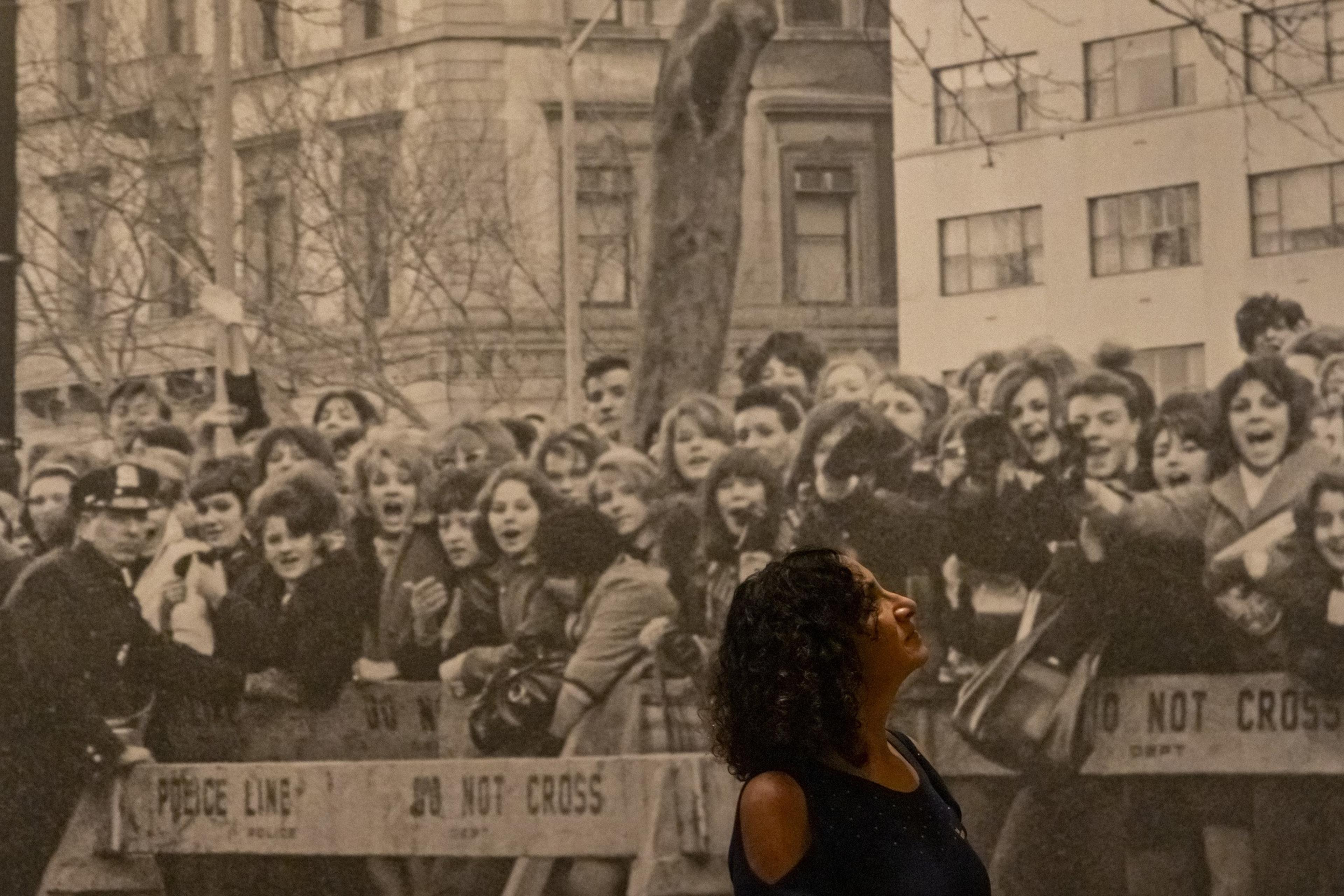 A woman in a dark top stands in front of a large black-and-white photo of a historical crowd scene, including a police officer and excited onlookers.