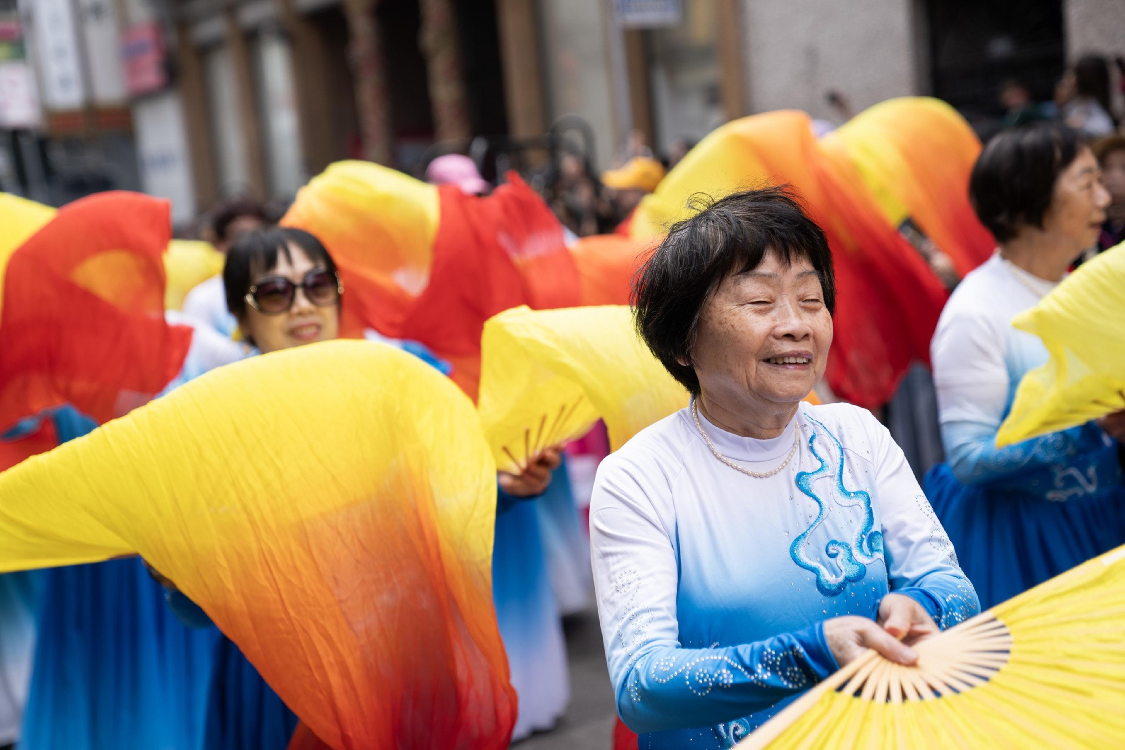 Older women wearing blue and white dresses dance outdoors, smiling and holding large yellow and red gradient fans during a cultural celebration.