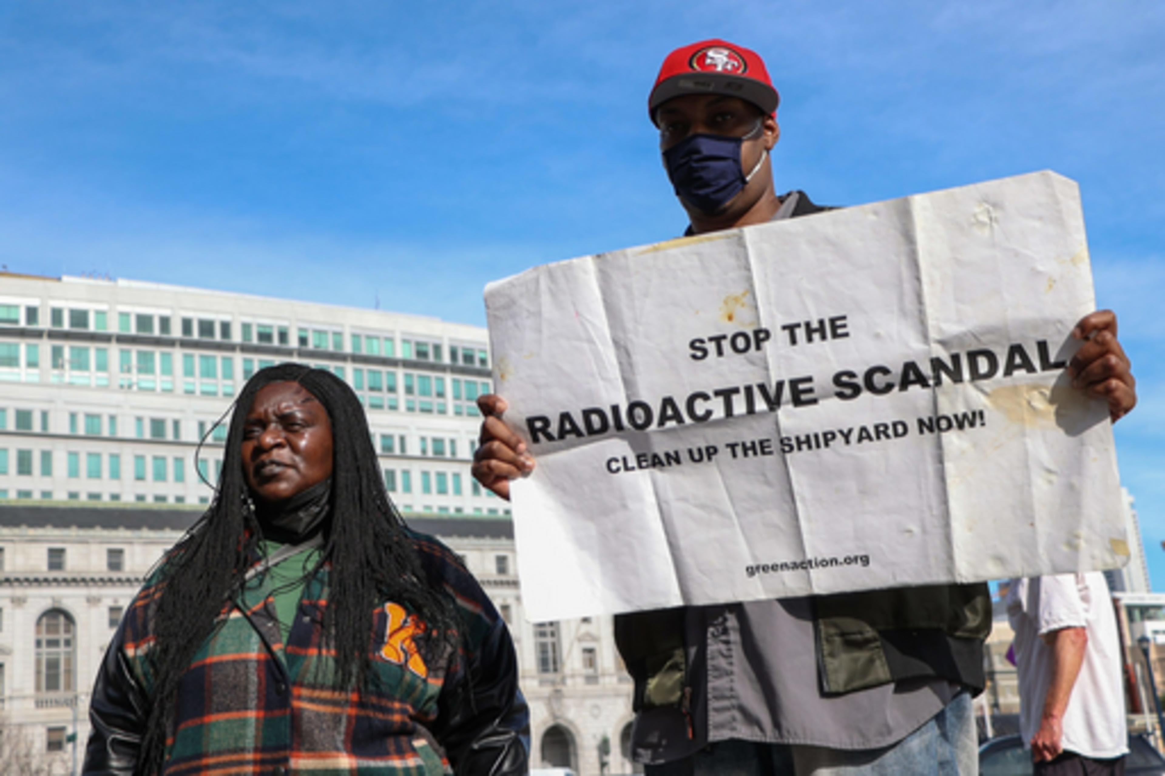 A man wearing a face mask and red cap holds a sign reading "Stop the radioactive scandal. Clean up the shipyard now!" next to a woman.
