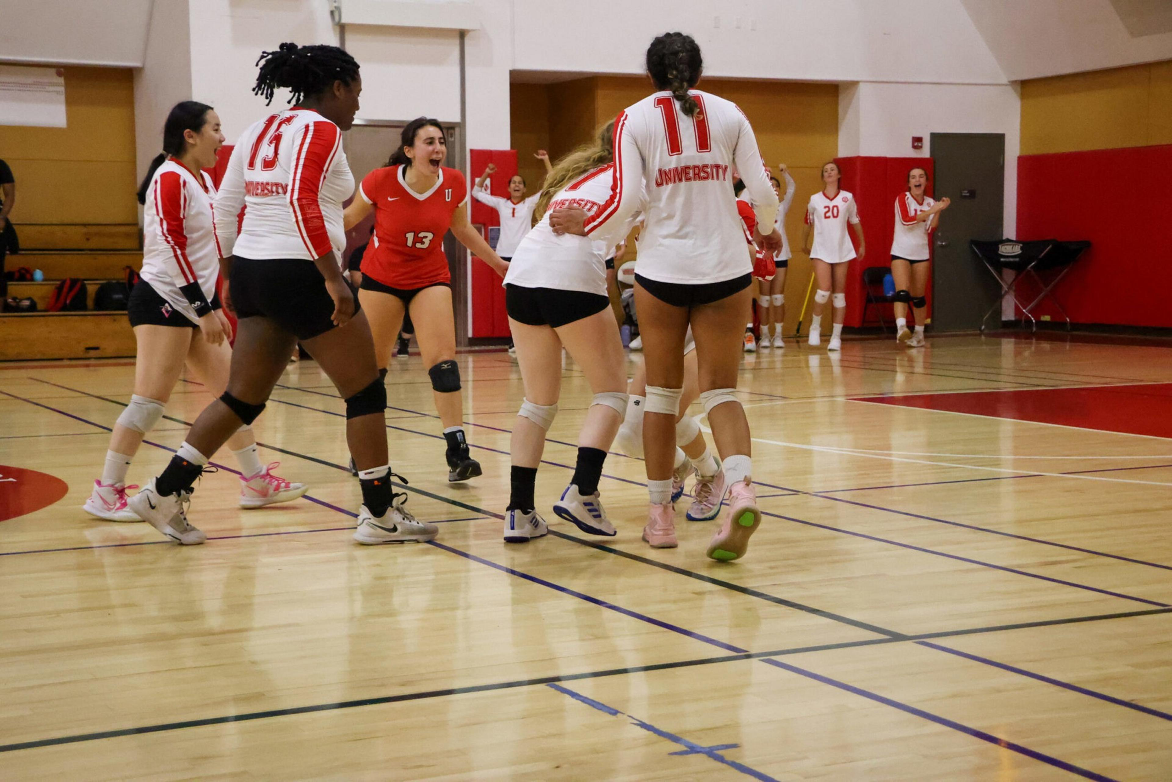 A women's volleyball team wearing white and red uniforms celebrates on an indoor court, showing excitement and joy together.