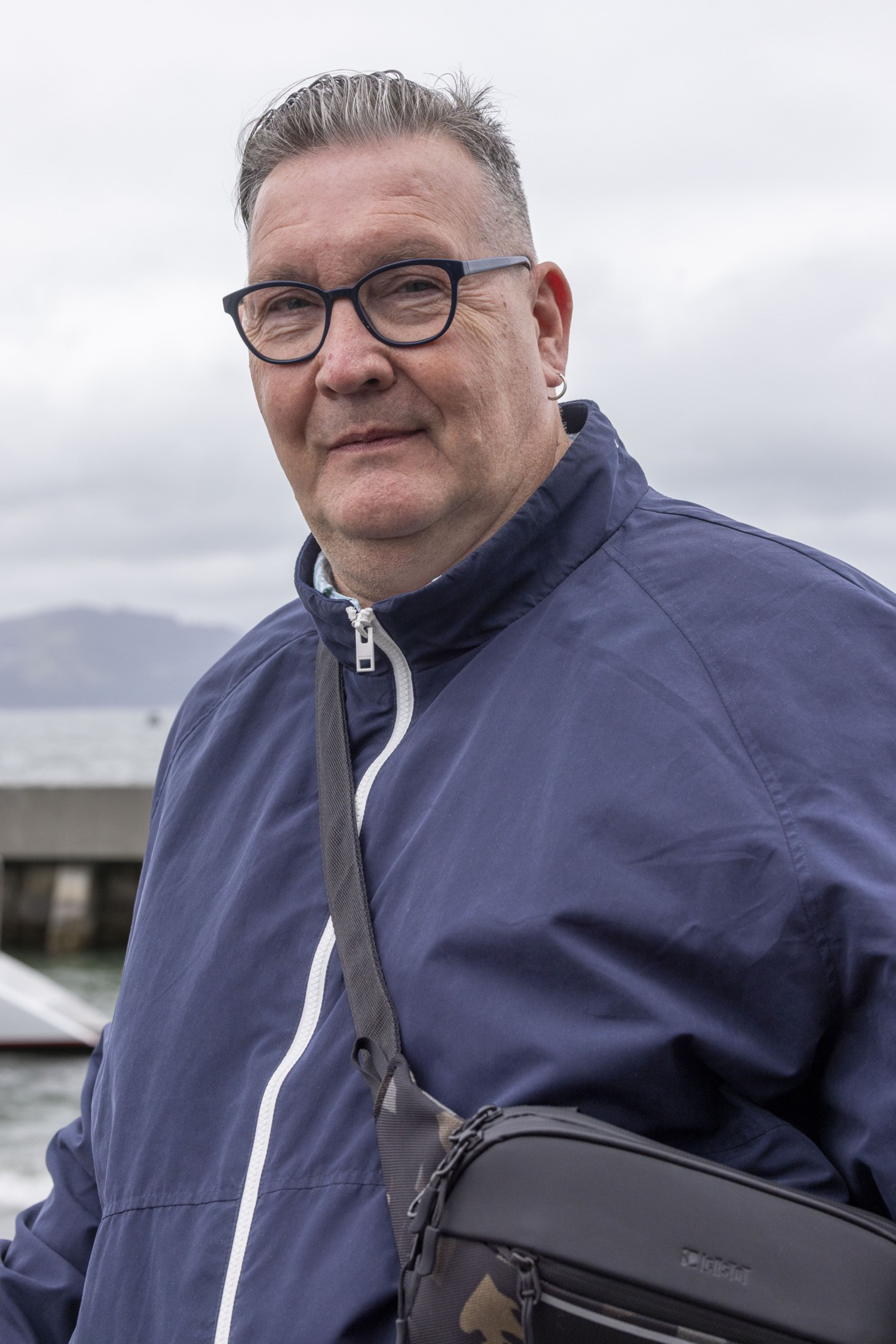 A man wearing glasses and a blue jacket stands outdoors, carrying a black crossbody bag. The background is cloudy with water and a mountain visible.