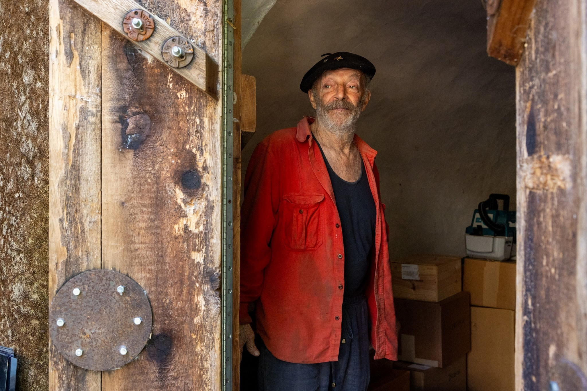 An older man with a beard, wearing a black beret, red shirt, and dark tank top, stands inside a rustic wooden doorway with boxes behind him.