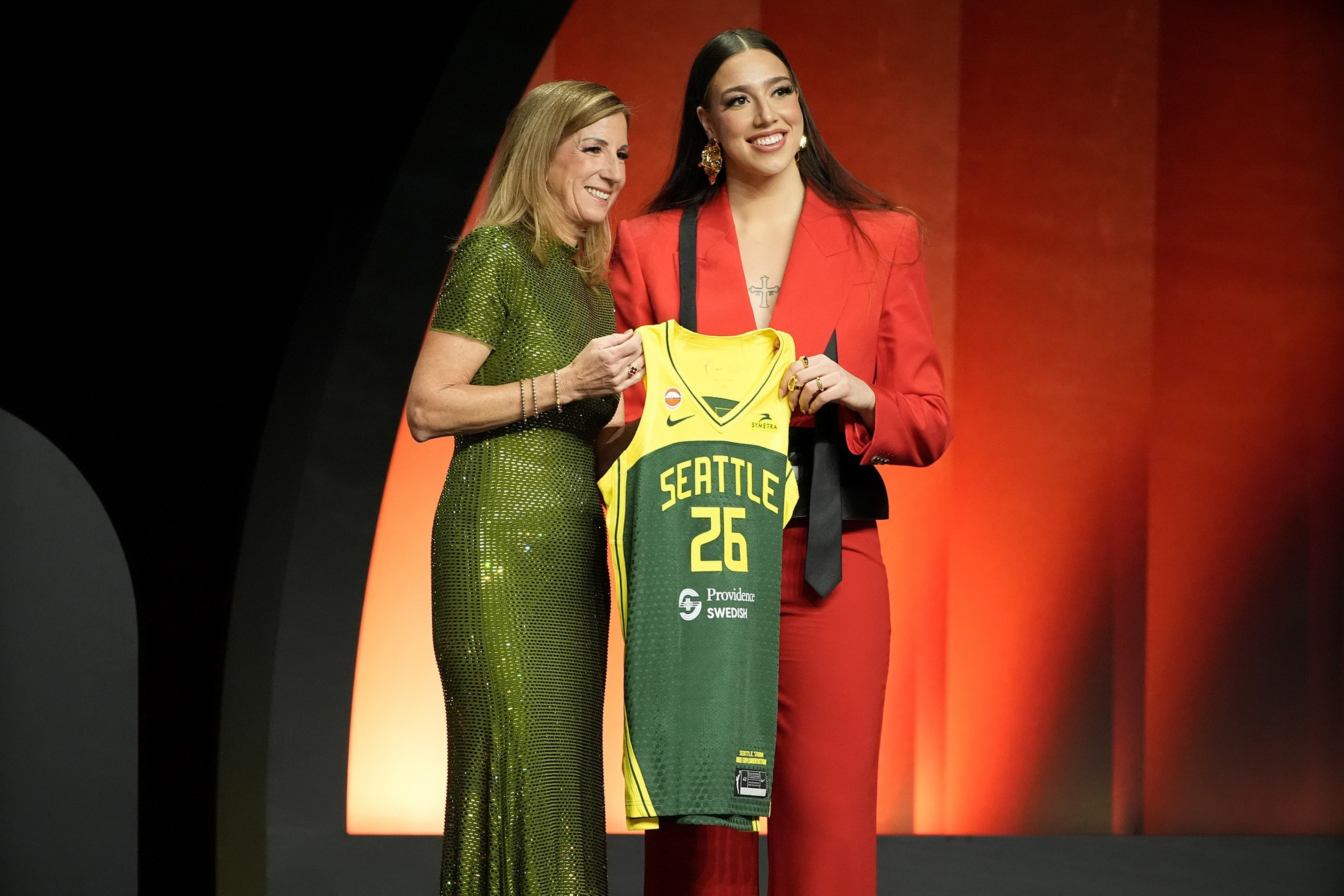 A woman in a green dress and a woman in a red suit smile while holding up a Seattle Storm basketball jersey with the number 26.