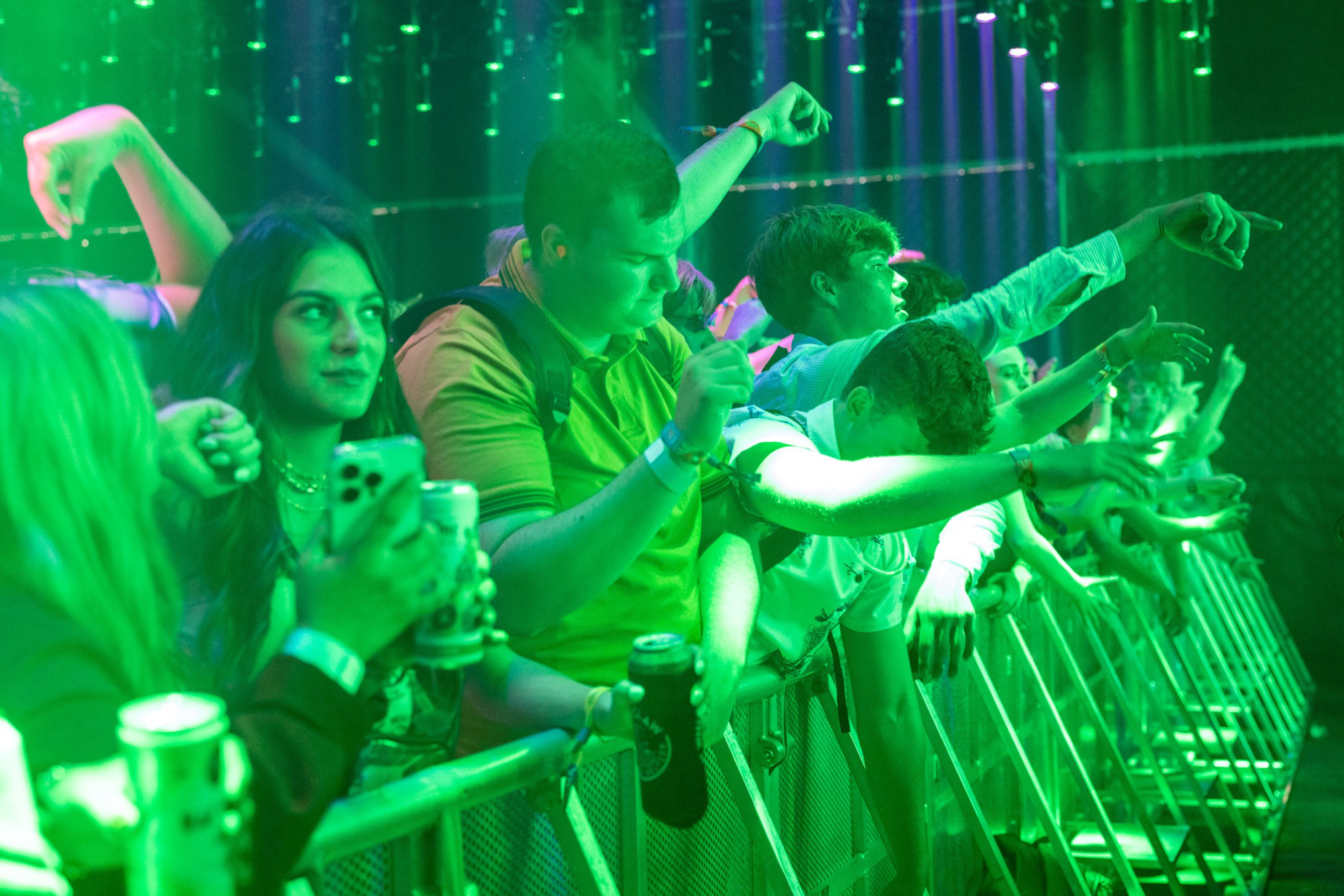 A crowd of young people at a concert leans against a barrier, raising their arms and holding drinks under green stage lighting.