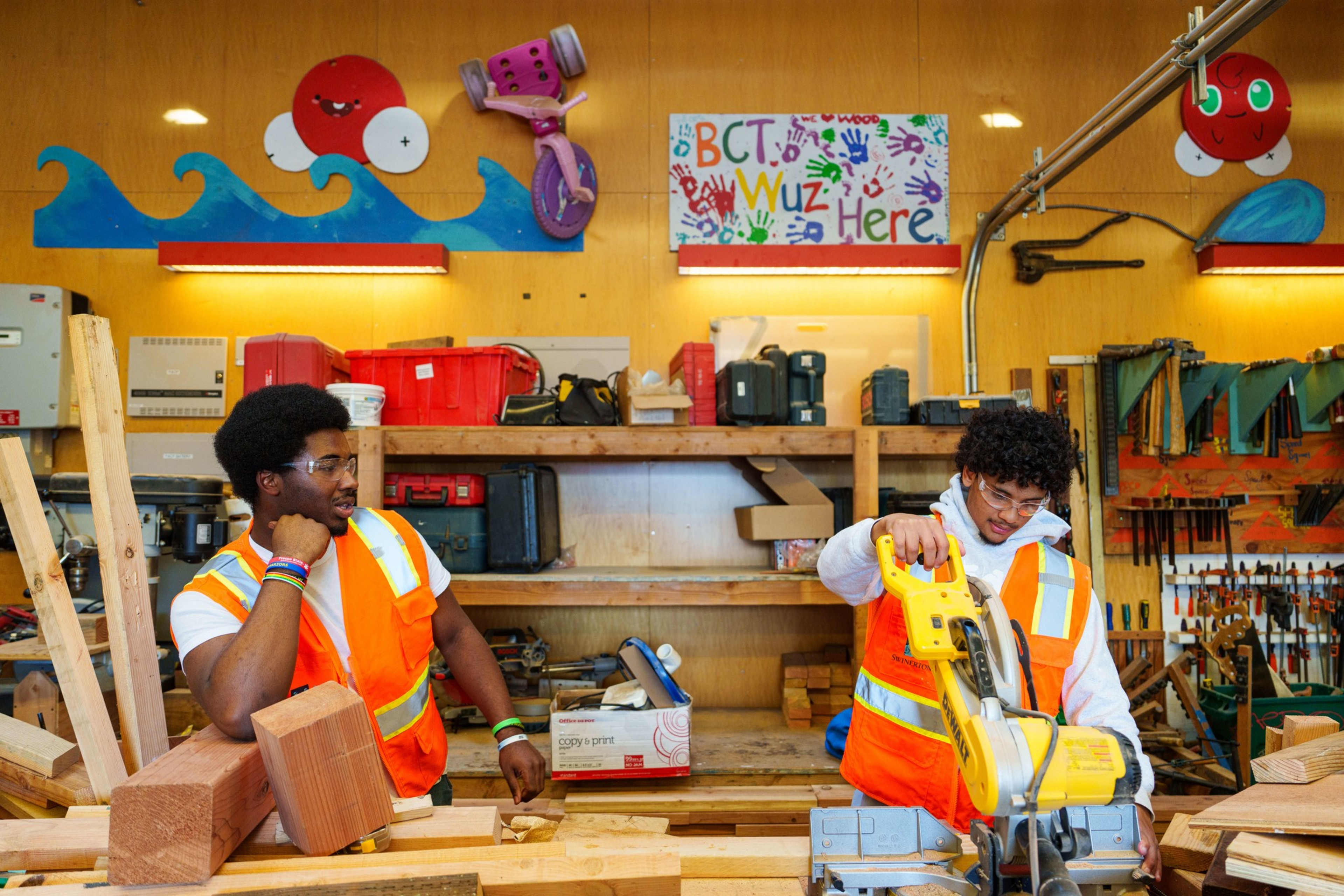 Two young men in orange safety vests work in a woodshop surrounded by wooden planks, tools, and colorful wall decorations with waves and a sun.