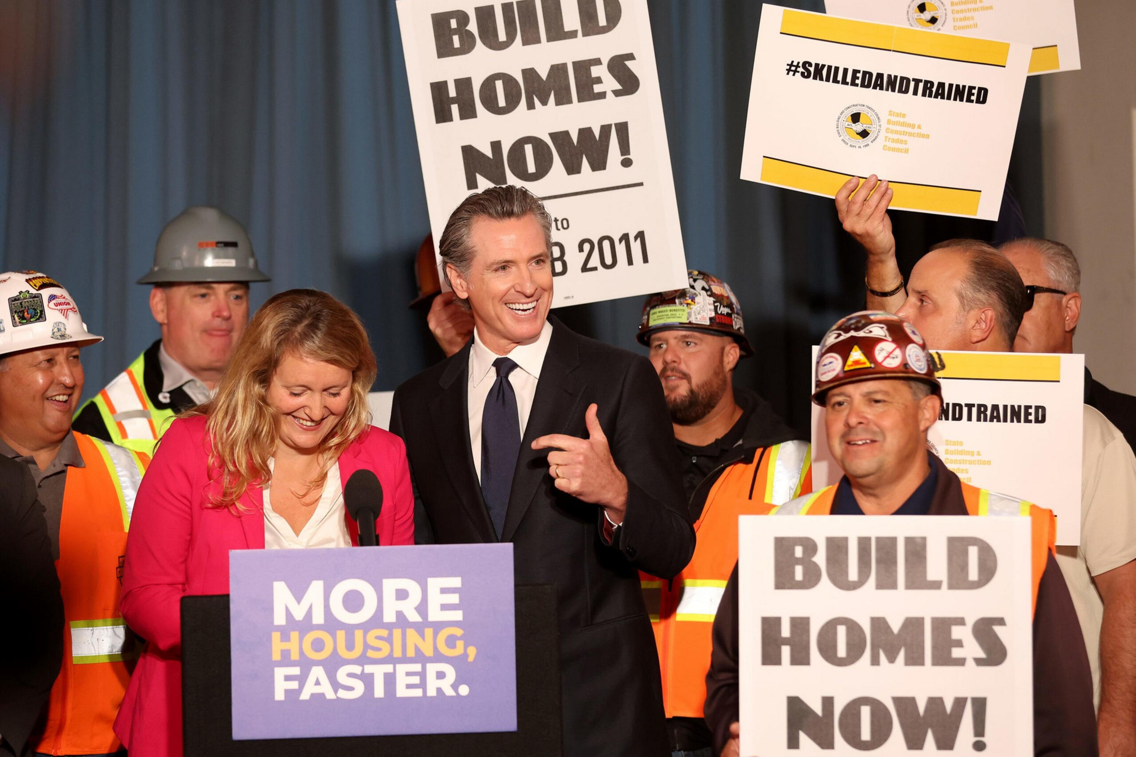 A group of people, including construction workers and two speakers at a podium, hold signs urging "Build Homes Now!" and "More Housing, Faster."