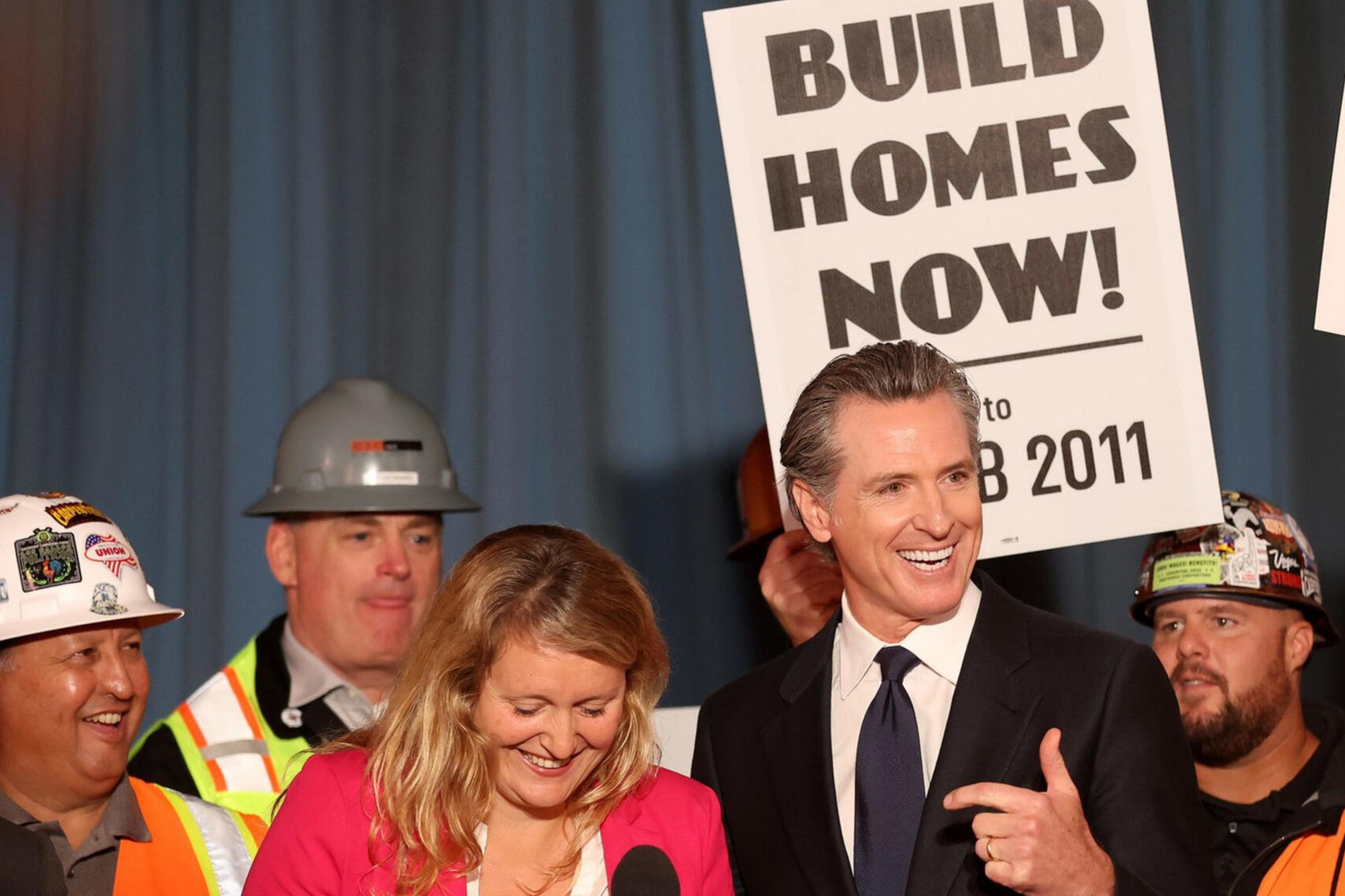 A group of people, including construction workers and two speakers at a podium, hold signs urging "Build Homes Now!" and "More Housing, Faster."