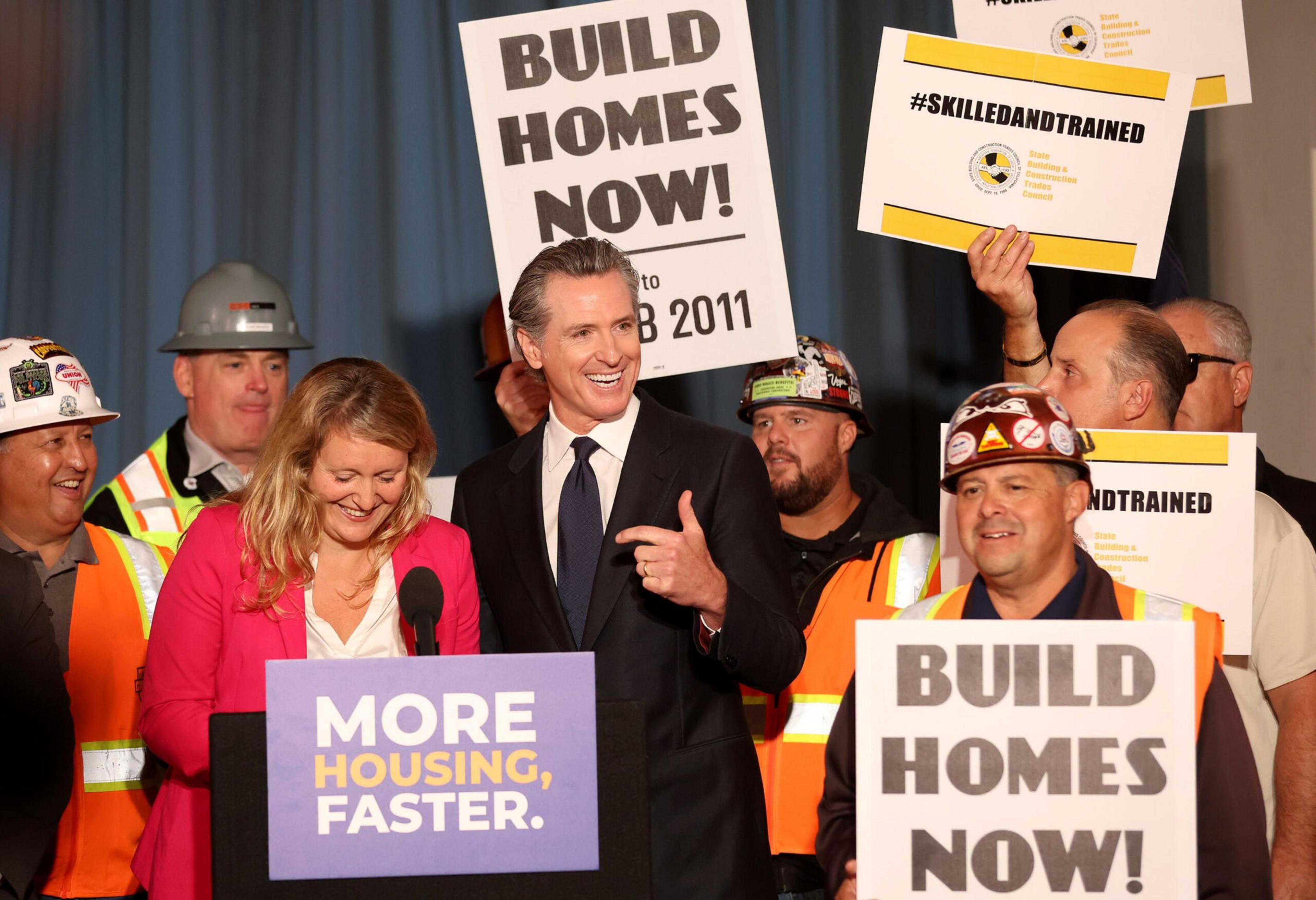 A group of people, including construction workers and two speakers at a podium, hold signs urging "Build Homes Now!" and "More Housing, Faster."