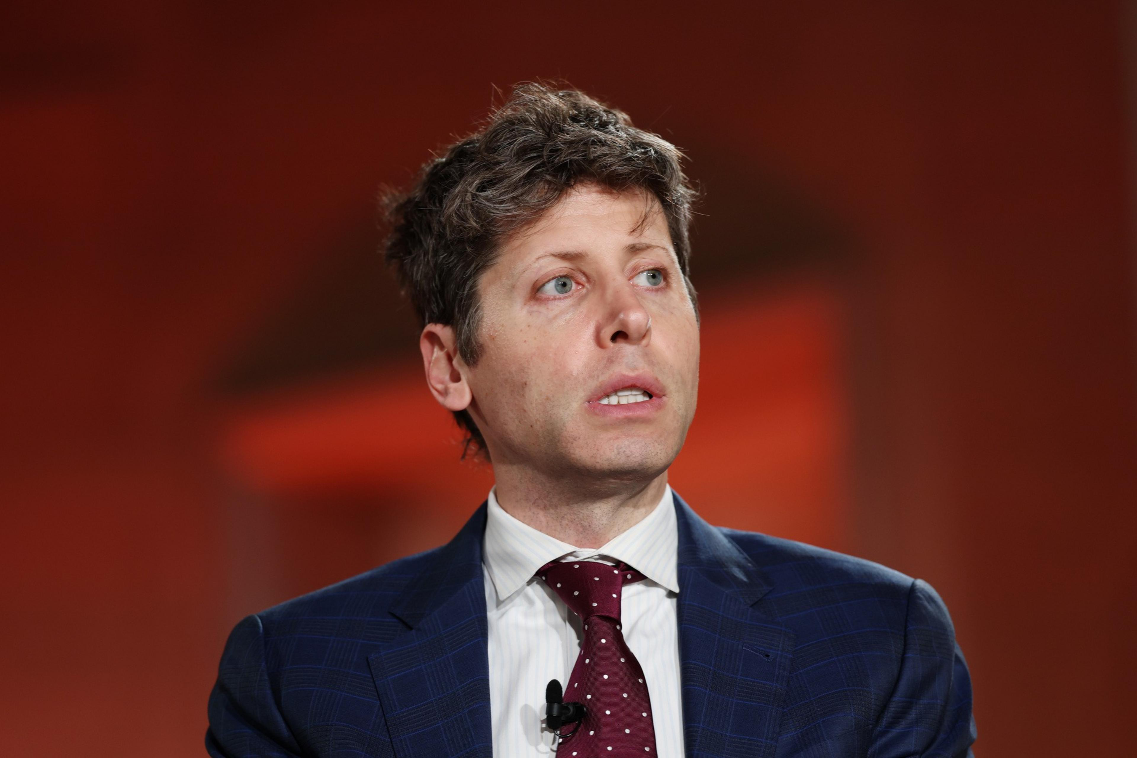 A man with short, curly brown hair wears a blue checkered suit, white shirt, and maroon polka dot tie, looking slightly to the right against a blurred orange background.