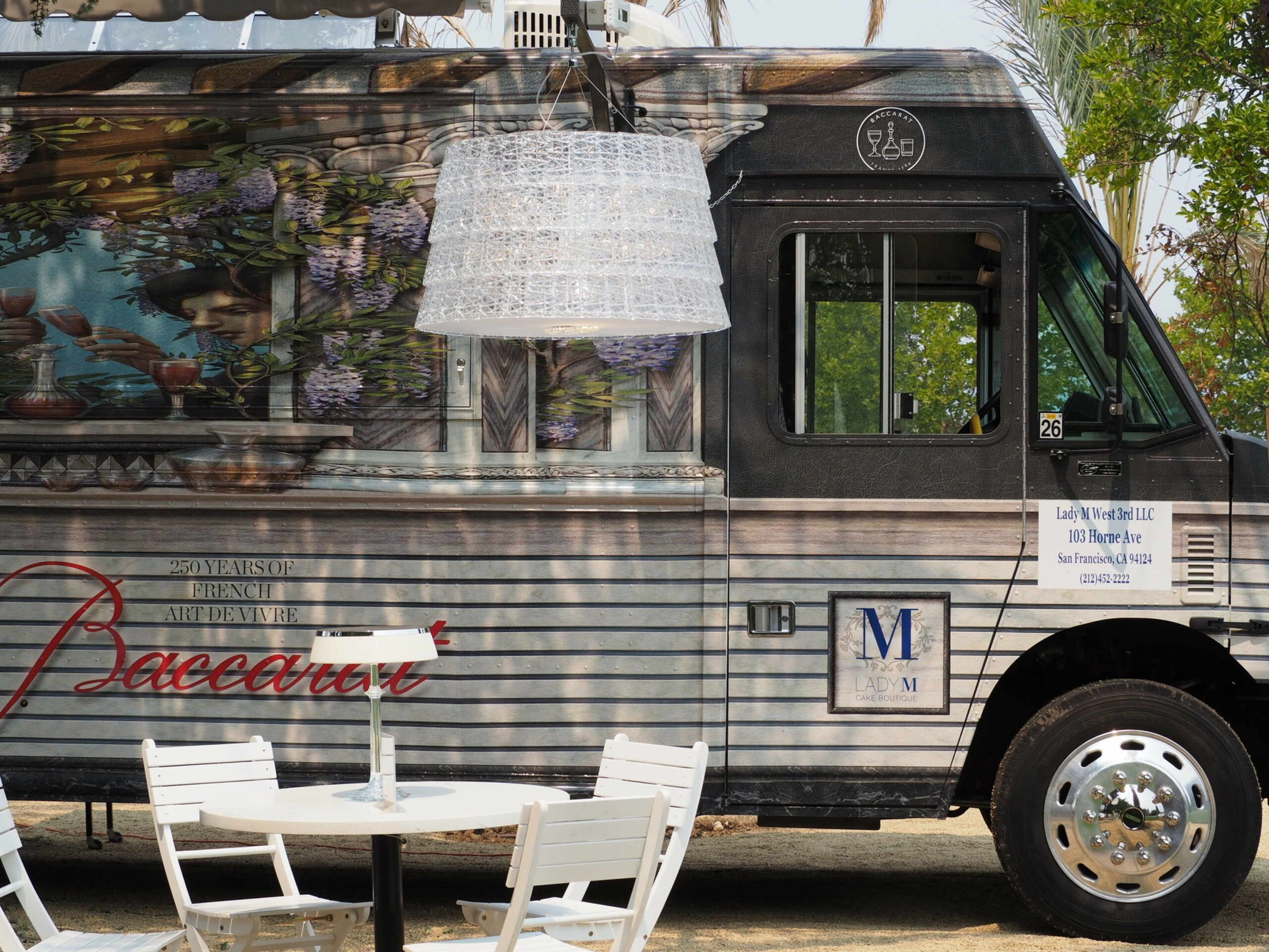 A black and gray food truck with "Baccarat" branding is parked beside a white outdoor table and chairs under a large hanging white lamp.