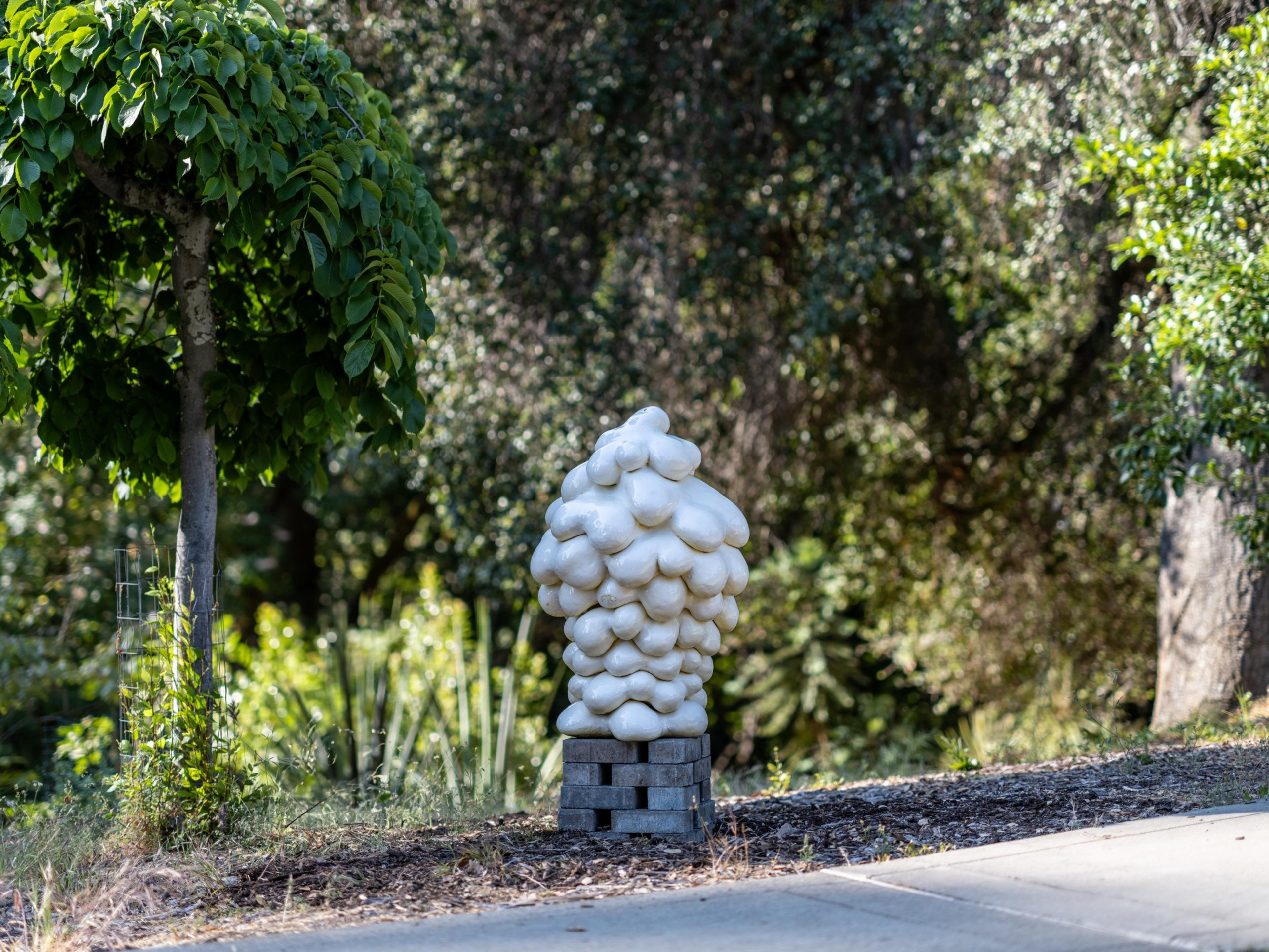 A cluster-like white sculpture sits on a platform beside a tree with dense green leaves, surrounded by lush greenery and a pathway.