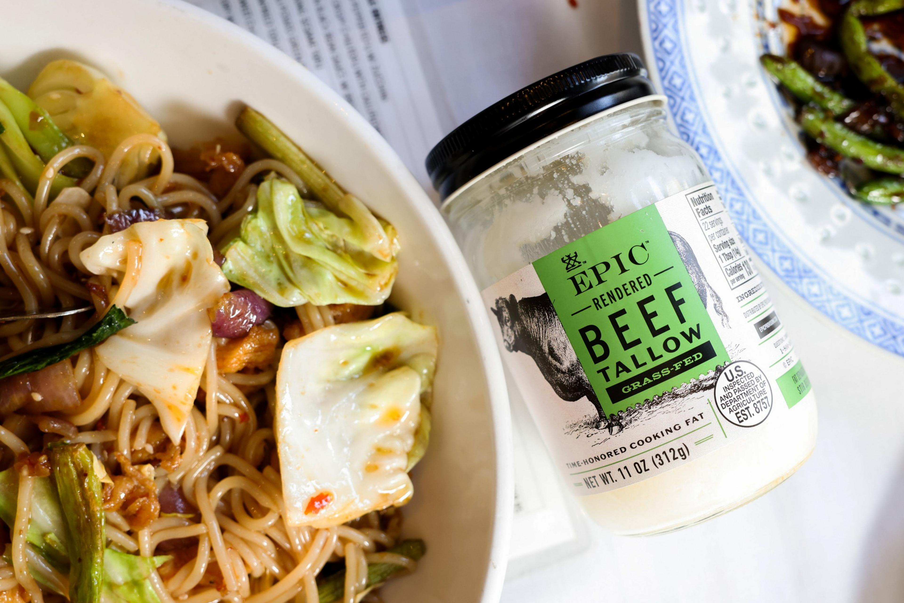 The image shows a bowl of noodles with vegetables. Next to it is a jar of Epic rendered beef tallow. A blue-edged plate with some food is partially visible.