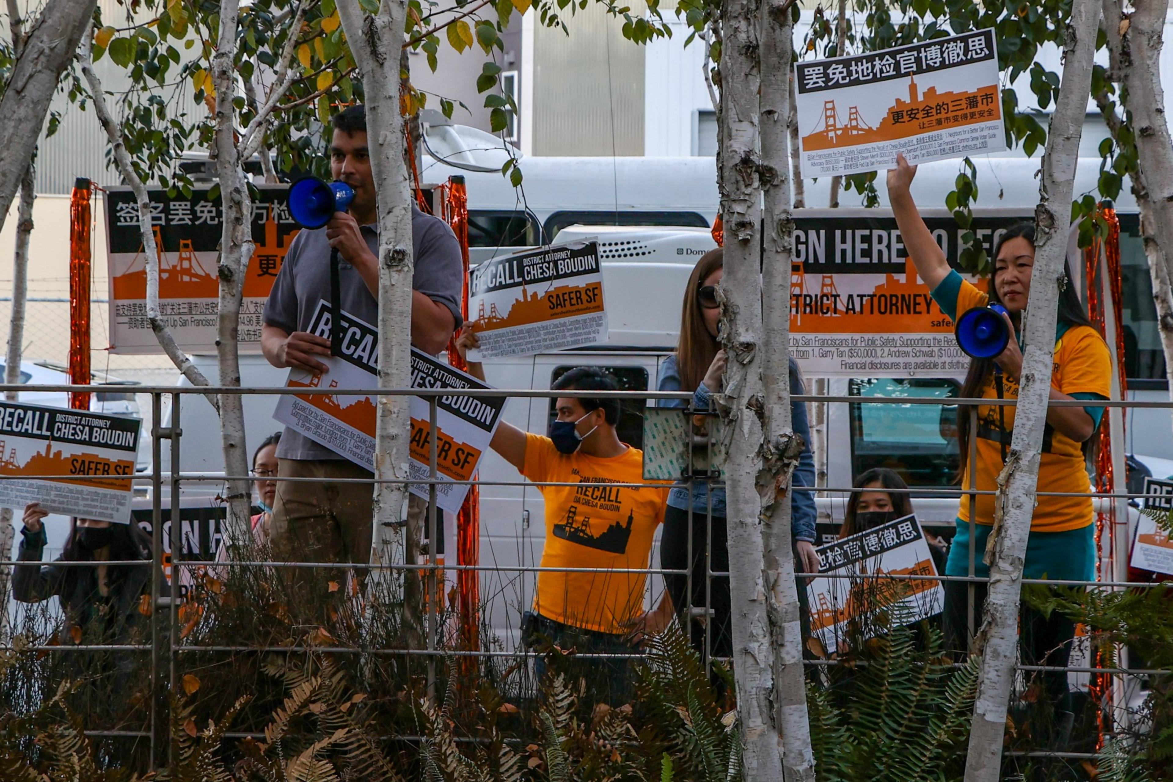 Several protesters hold signs calling to recall Chesa Boudin, some using megaphones, standing behind a metal fence with trees partially obscuring them.