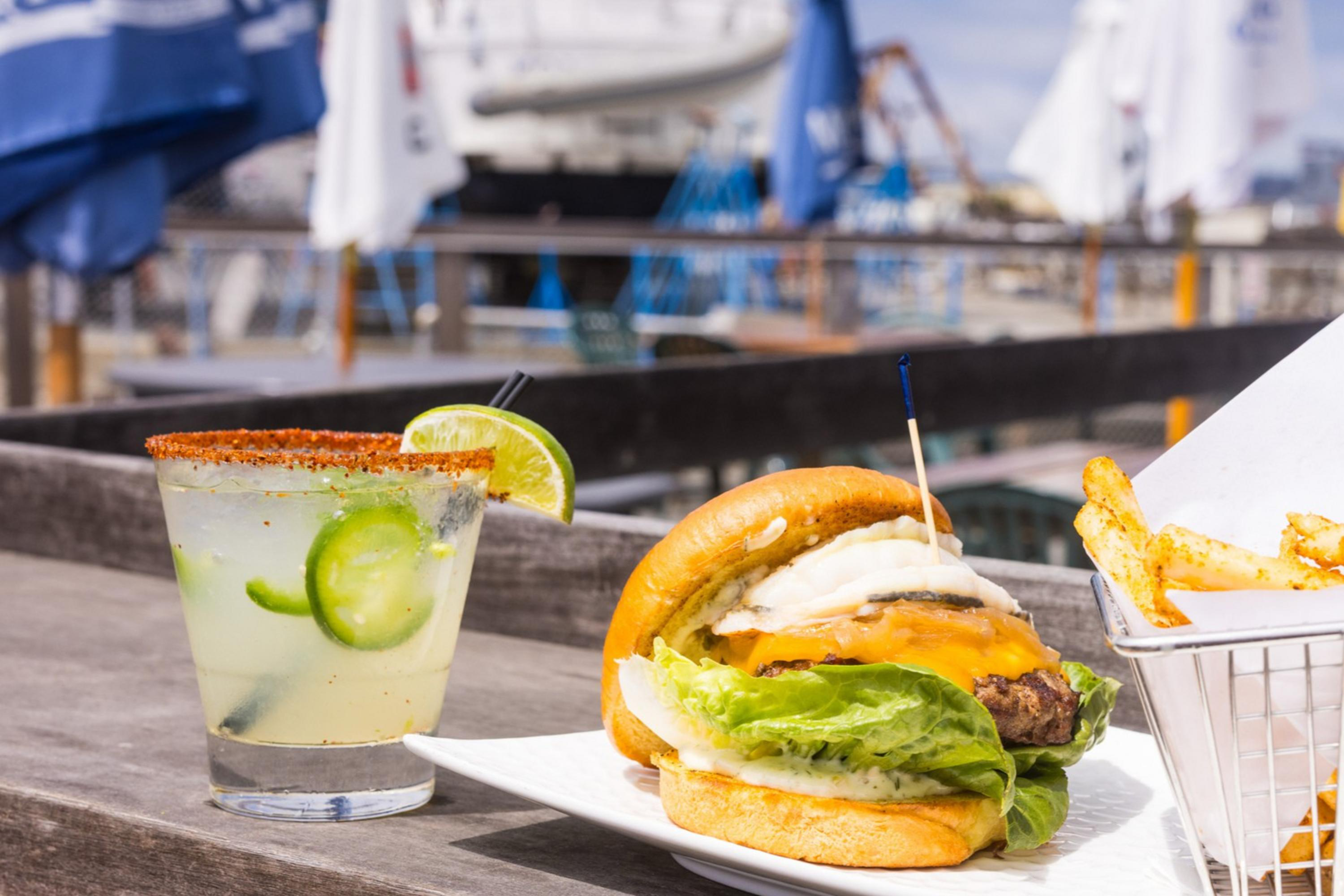 A cheeseburger with lettuce and onion sits on a plate with a basket of fries. A cocktail garnished with lime and jalapeño is beside it; a boat and umbrellas appear in the background.