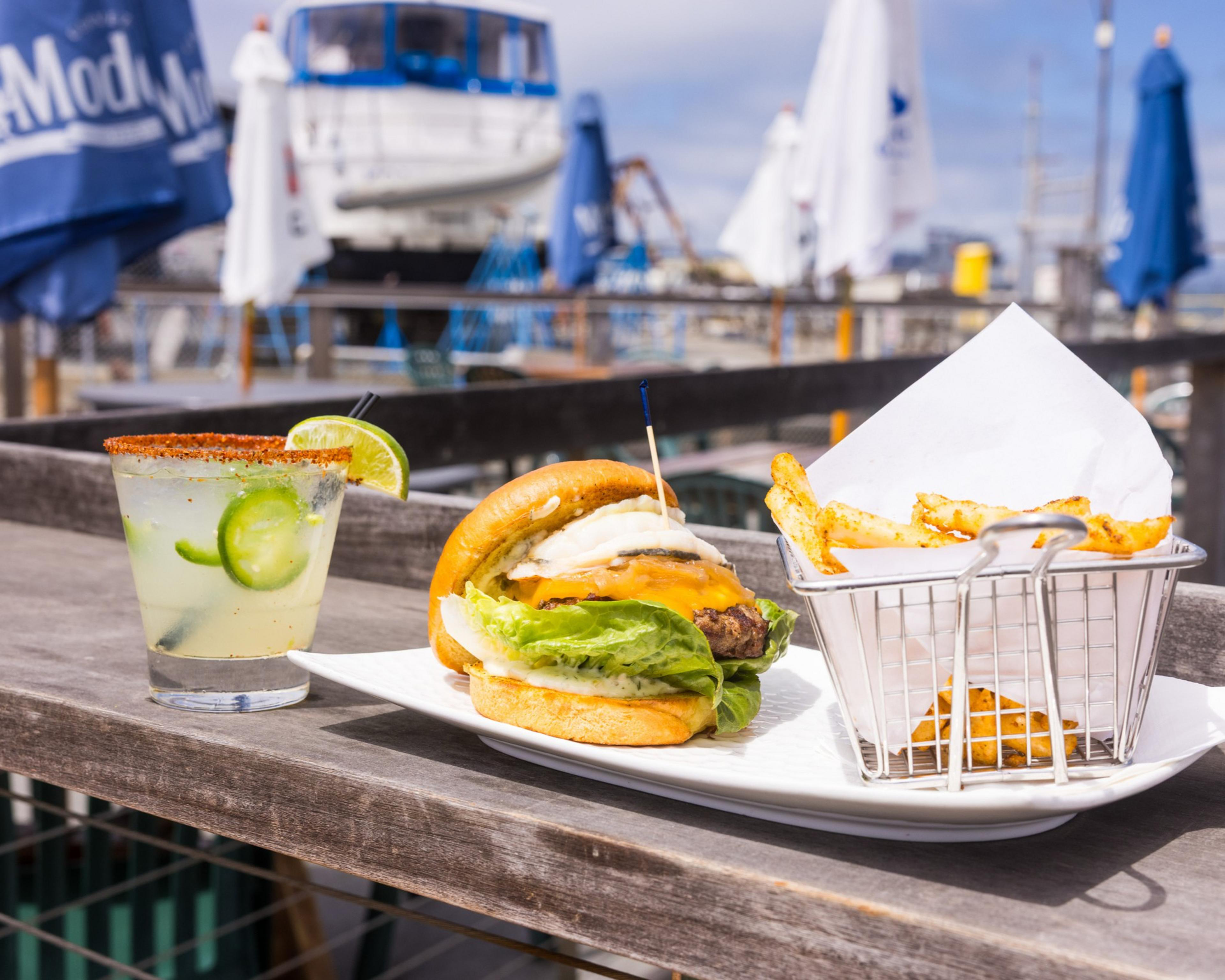 A cheeseburger with lettuce and onion sits on a plate with a basket of fries. A cocktail garnished with lime and jalapeño is beside it; a boat and umbrellas appear in the background.