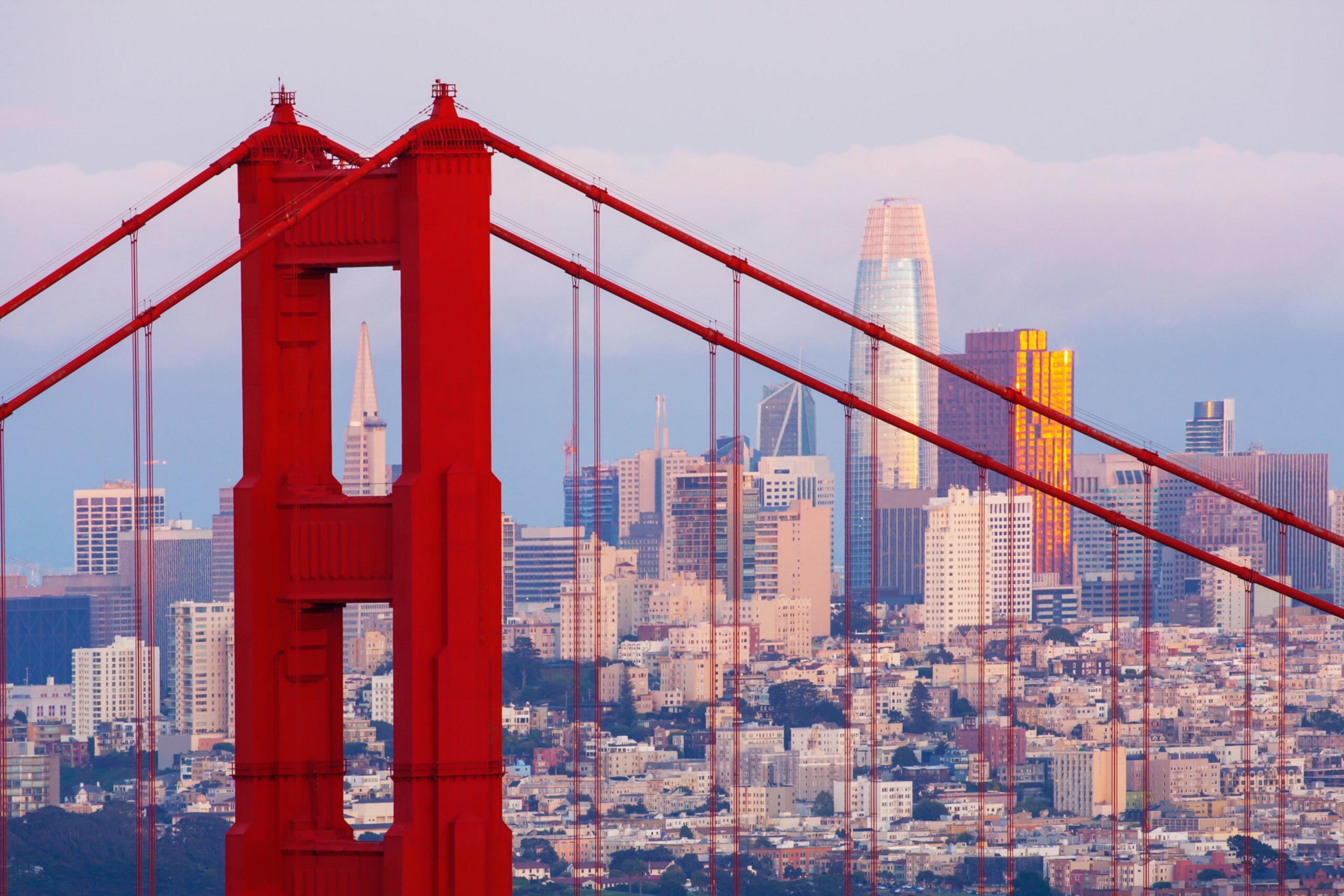 A close-up of the red Golden Gate Bridge towers with cables, set against the backdrop of San Francisco's city skyline under a partly cloudy sky.
