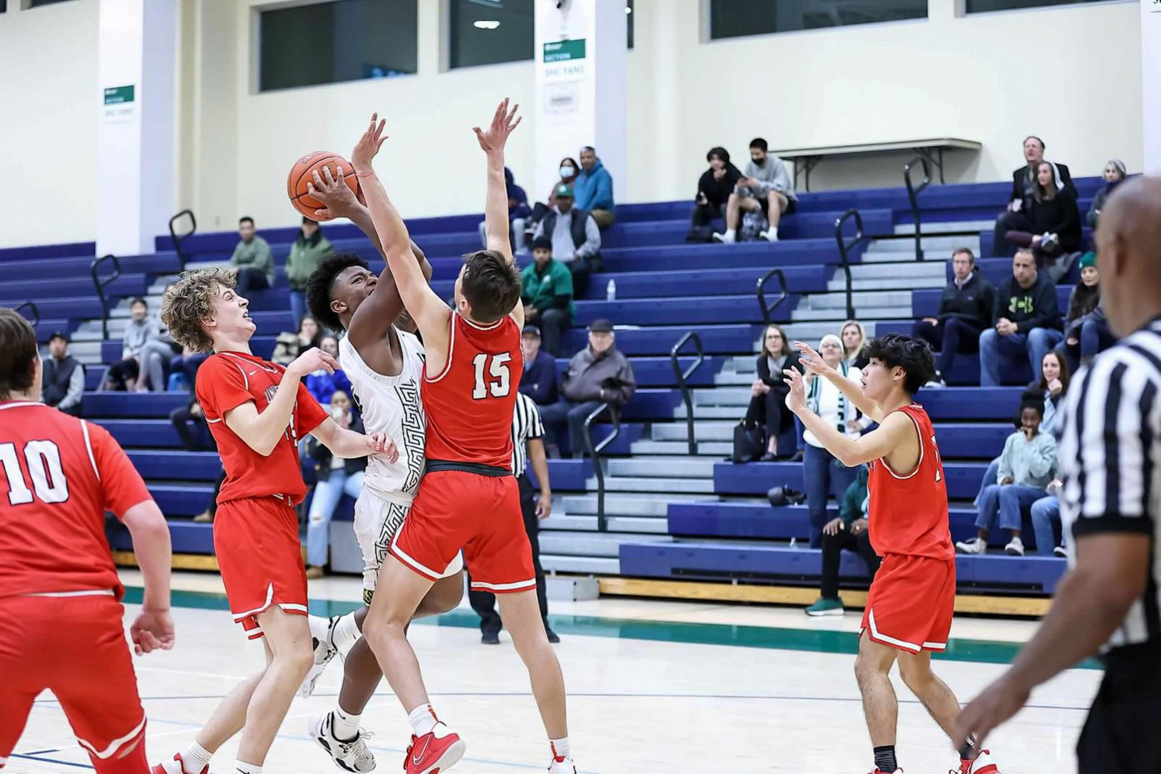 A basketball player in white attempts a shot while three players in red defend, with spectators watching from bleachers in the background.
