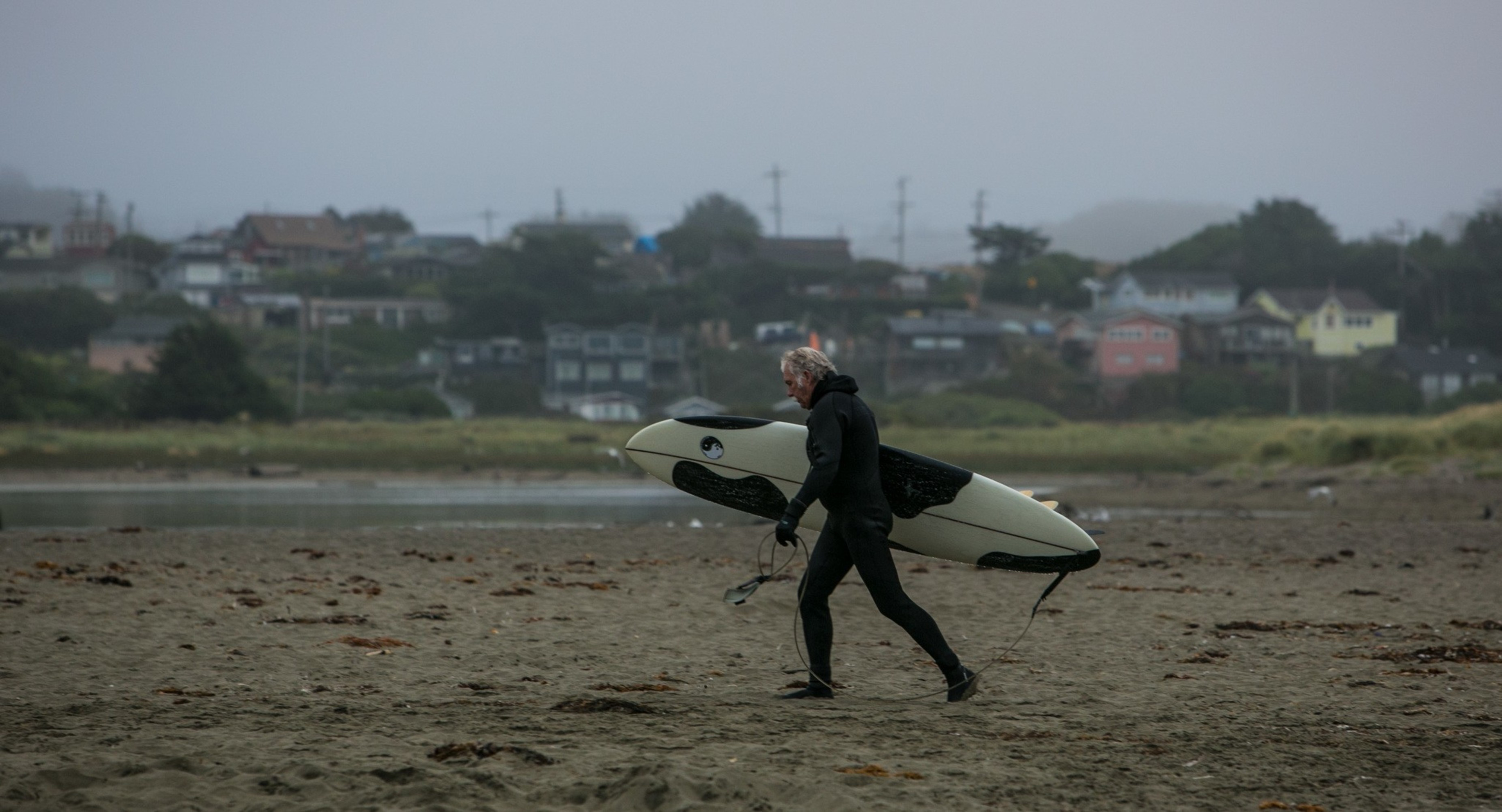 A person wearing a black wetsuit walks across a sandy beach carrying a white and black surfboard under a gray, foggy sky.