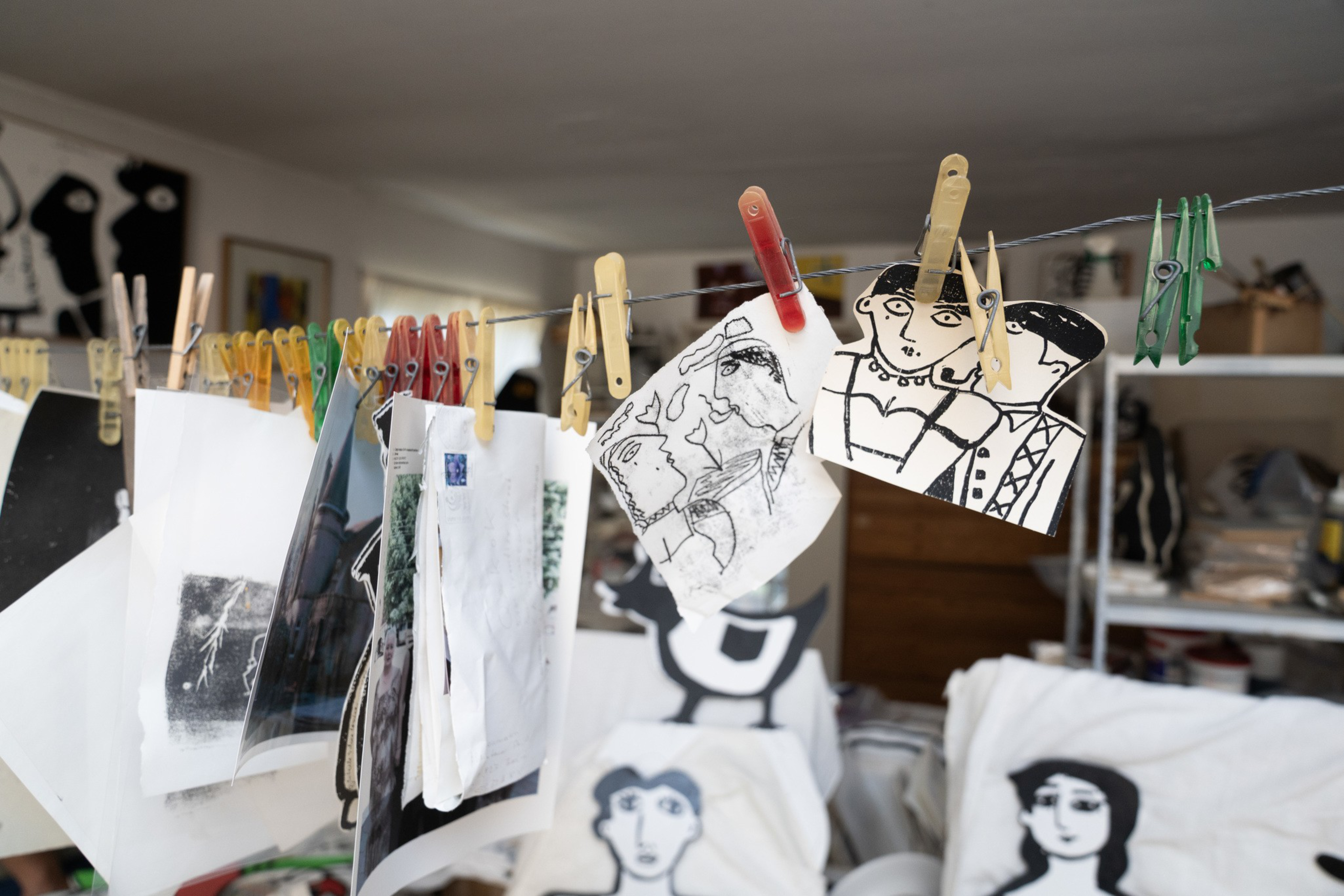 Black and white drawings of faces and figures hang from clothespins on a line in an art studio filled with similar artwork and art supplies.