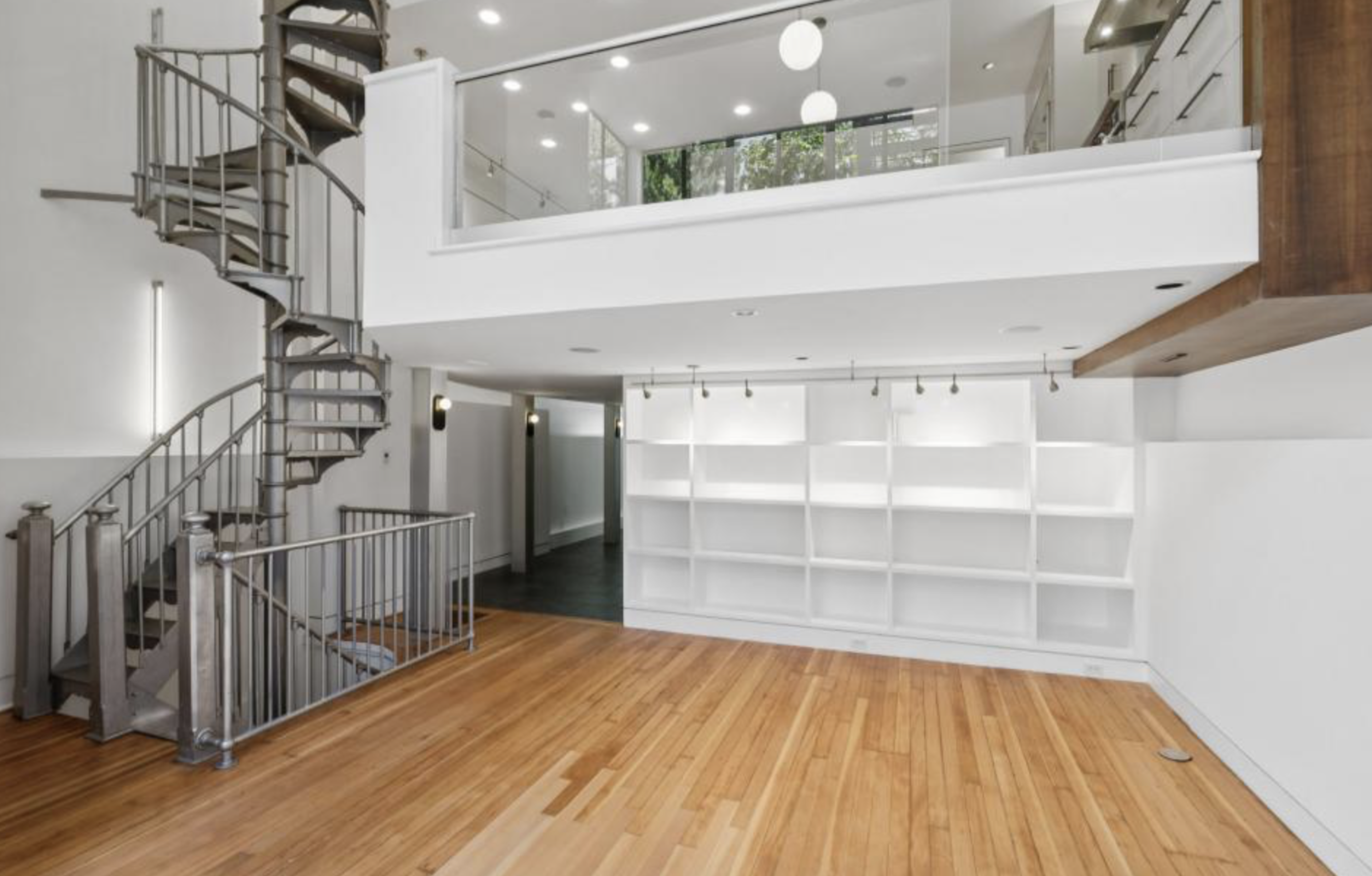 An open room with light wood flooring, white built-in shelves, a spiral staircase, glass railing, and a mezzanine level with large windows.