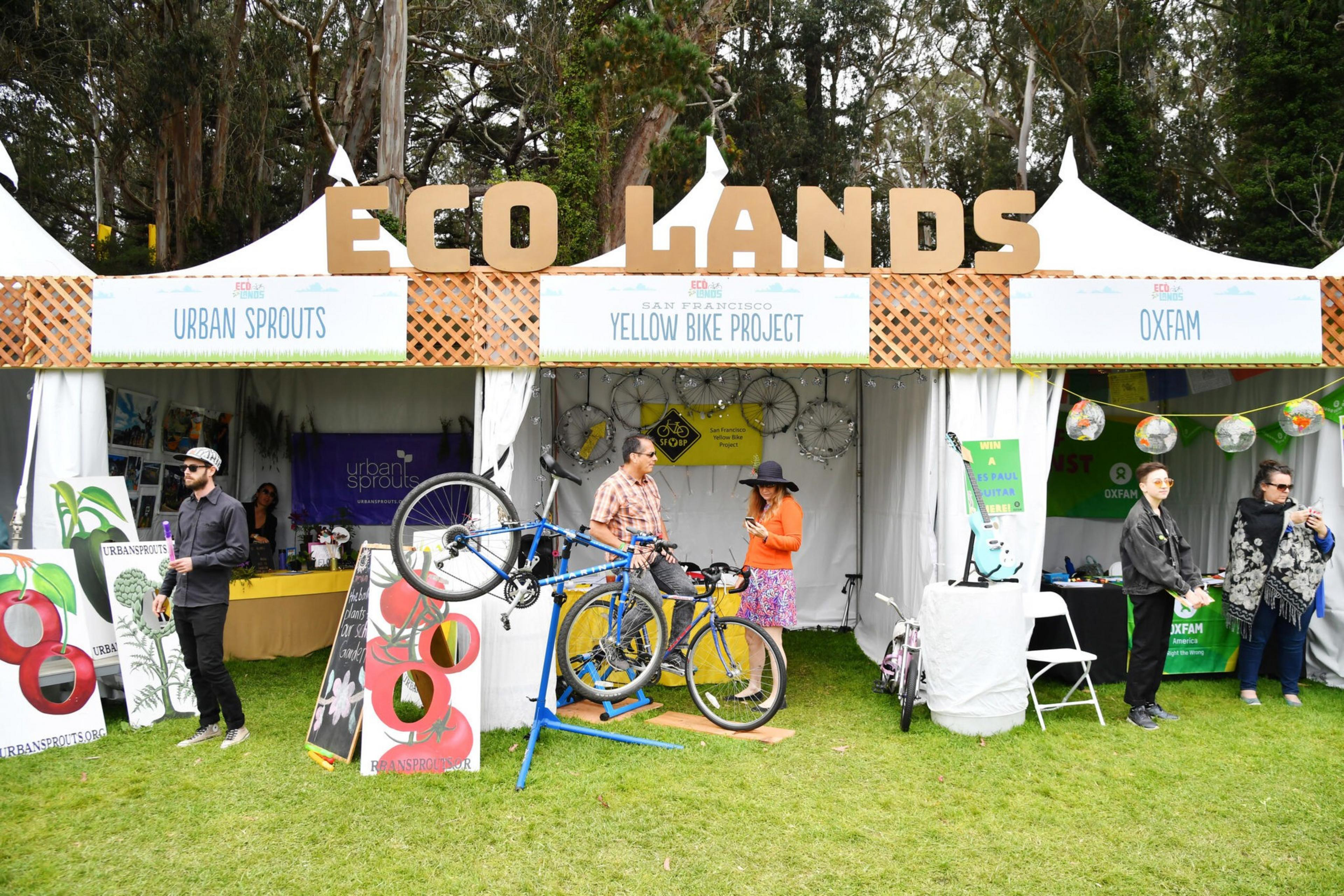 Three adjacent tents labeled Urban Sprouts, Yellow Bike Project, and Oxfam, with people interacting and a bike repair setup in an outdoor grassy area.
