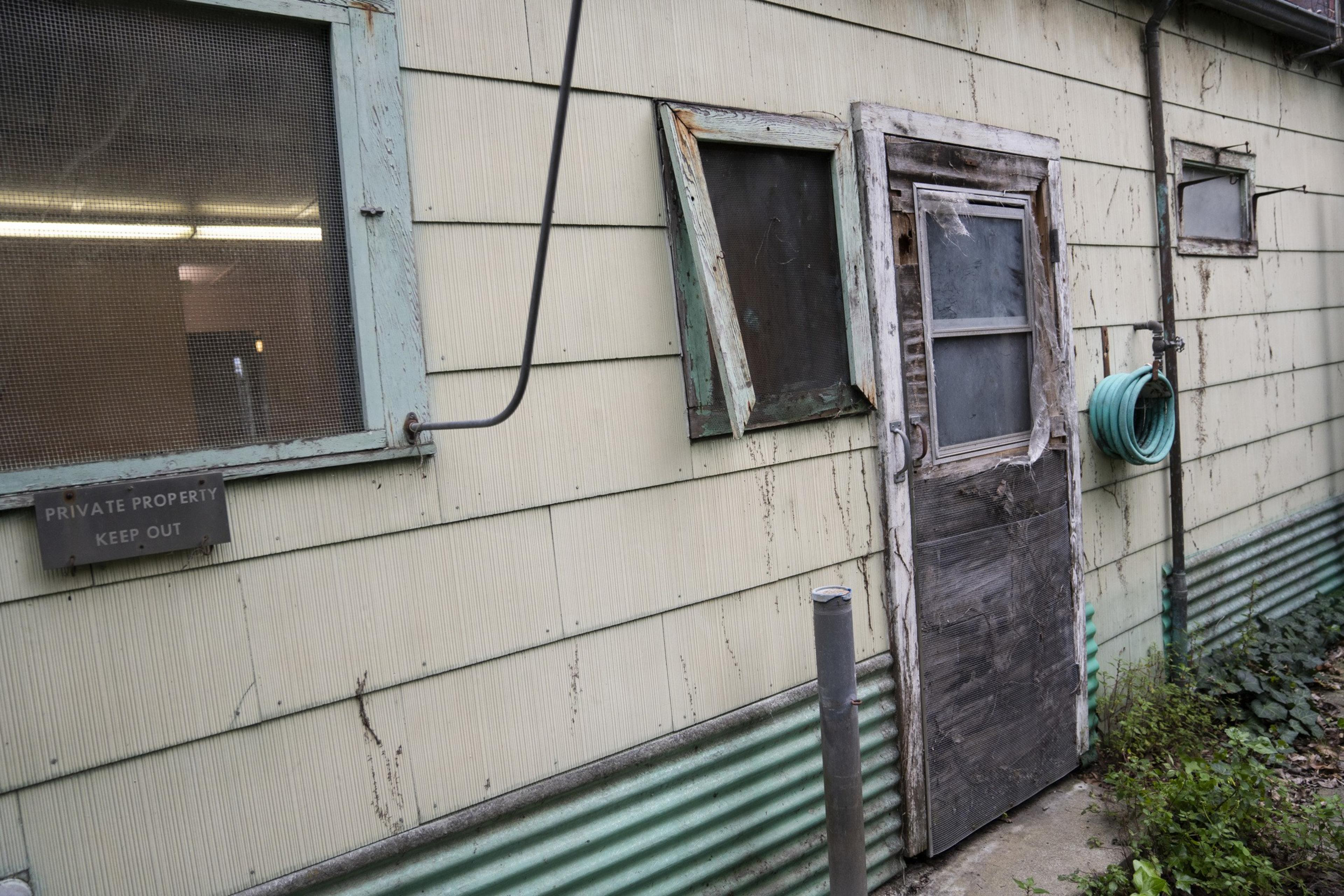 A weathered building with a worn door and windows, a &quot;Private Property Keep Out&quot; sign, and a green hose coiled on the exterior wall.