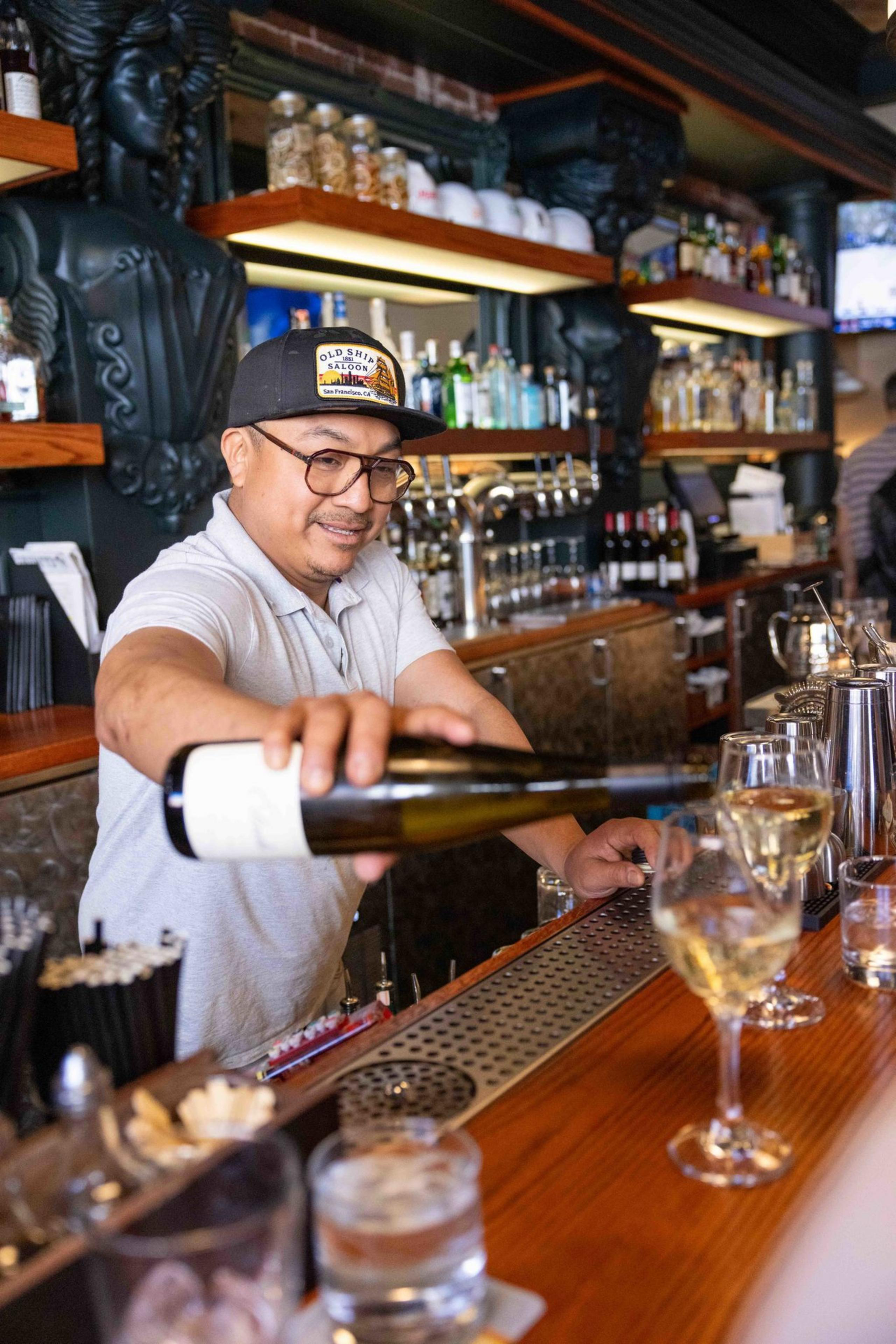 A bartender in a cap and glasses pours white wine into a glass at a bar. Shelves behind him hold bottles, and various drinks are on the counter.