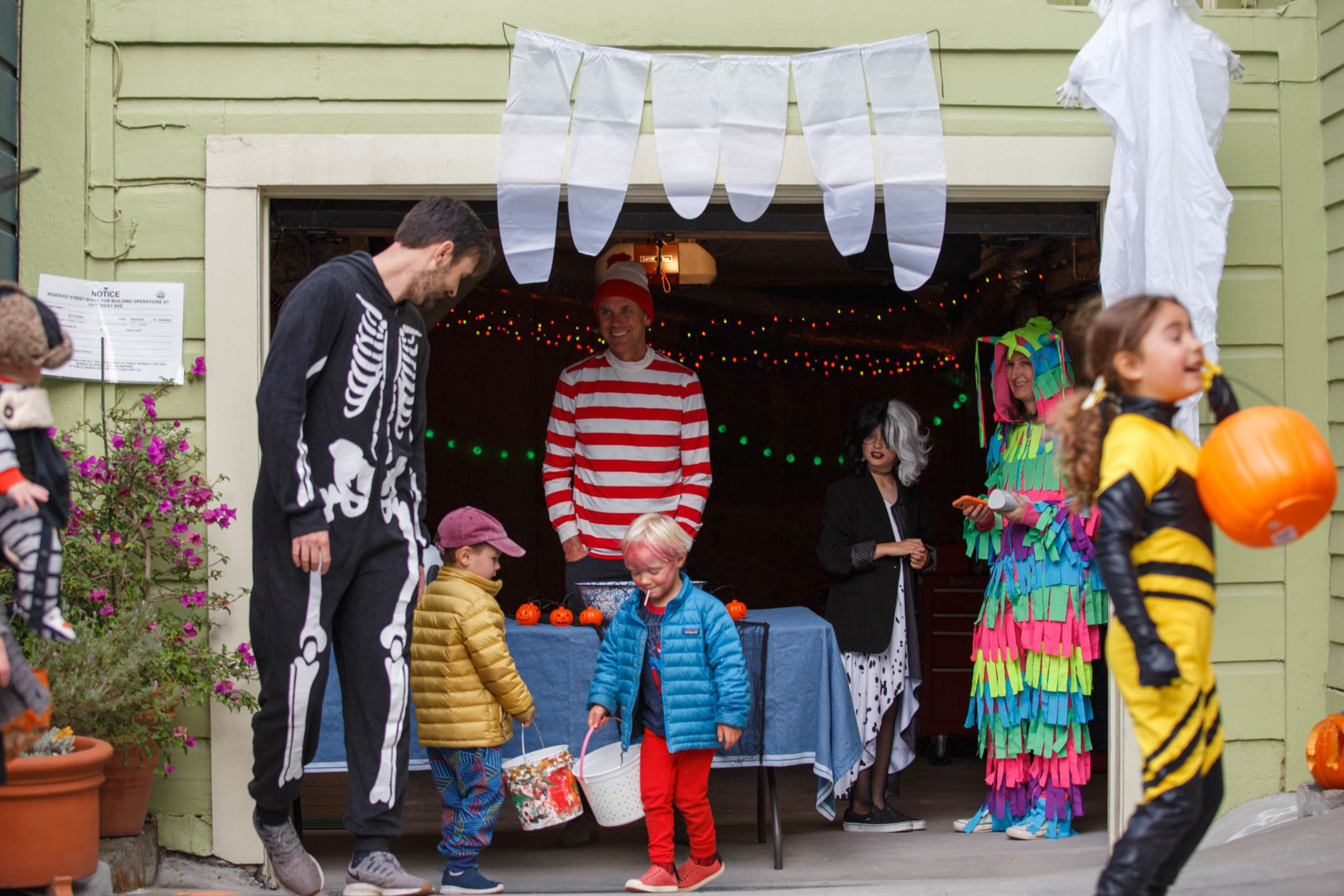 Children and adults in Halloween costumes, including a skeleton, Waldo, and a colorful piñata, gather outside a decorated garage for trick-or-treating.