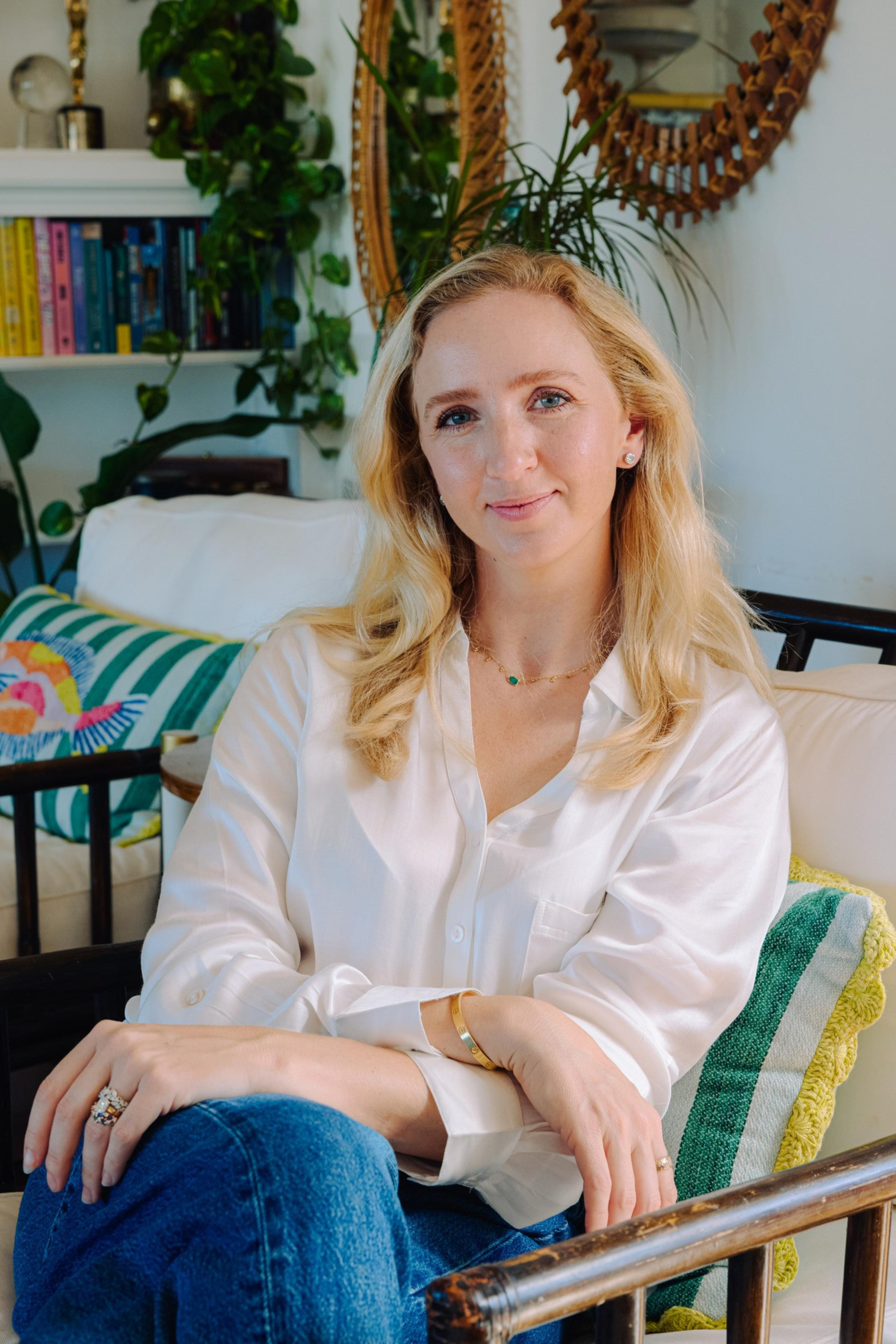 A woman with long blonde hair sits on a sofa, wearing a white shirt and blue jeans. The room has plants, colorful pillows, books, and ornate mirrors.
