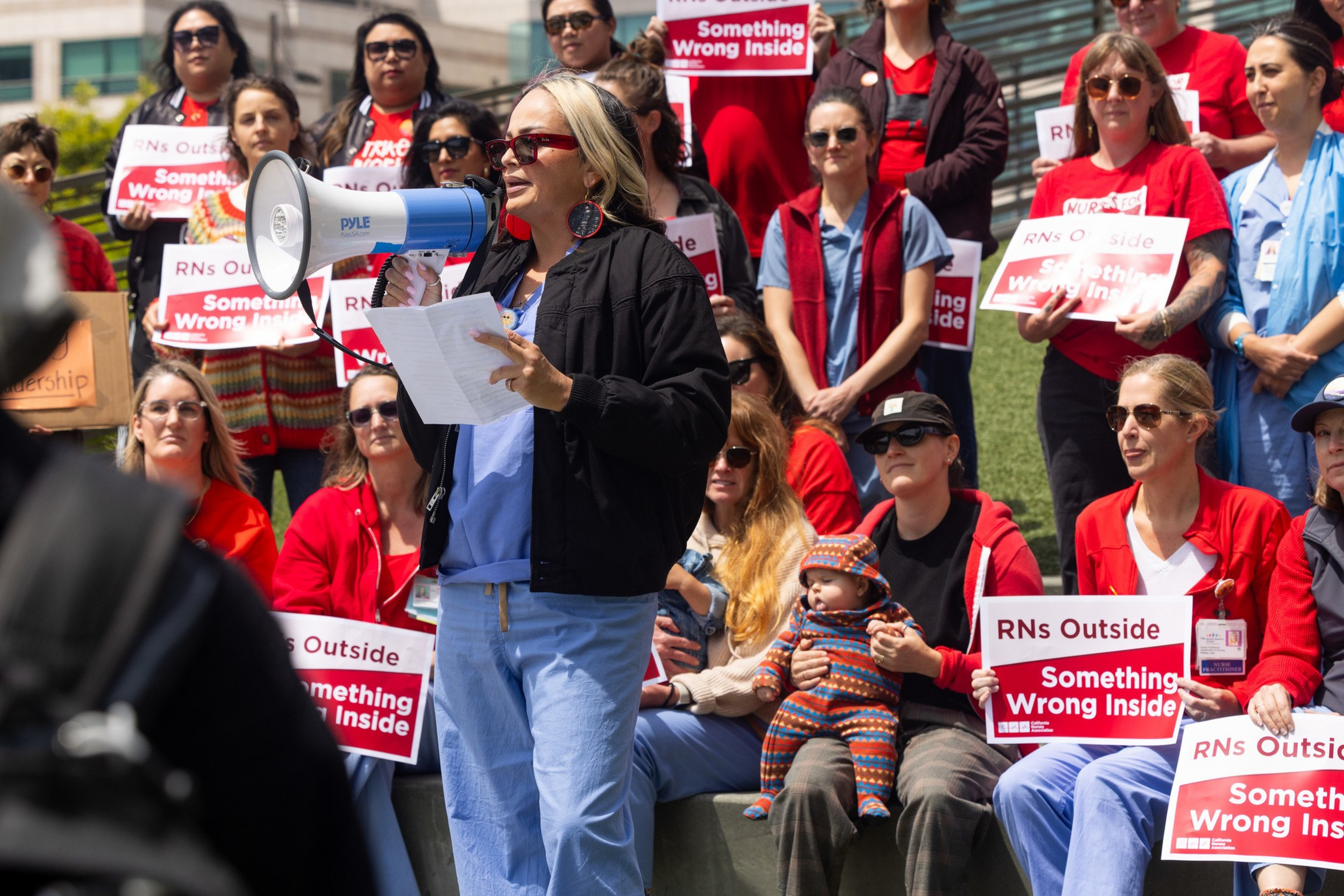 A woman speaks into a megaphone while a crowd, including seated people holding signs reading “RNs Outside Something Wrong Inside,” listens attentively.