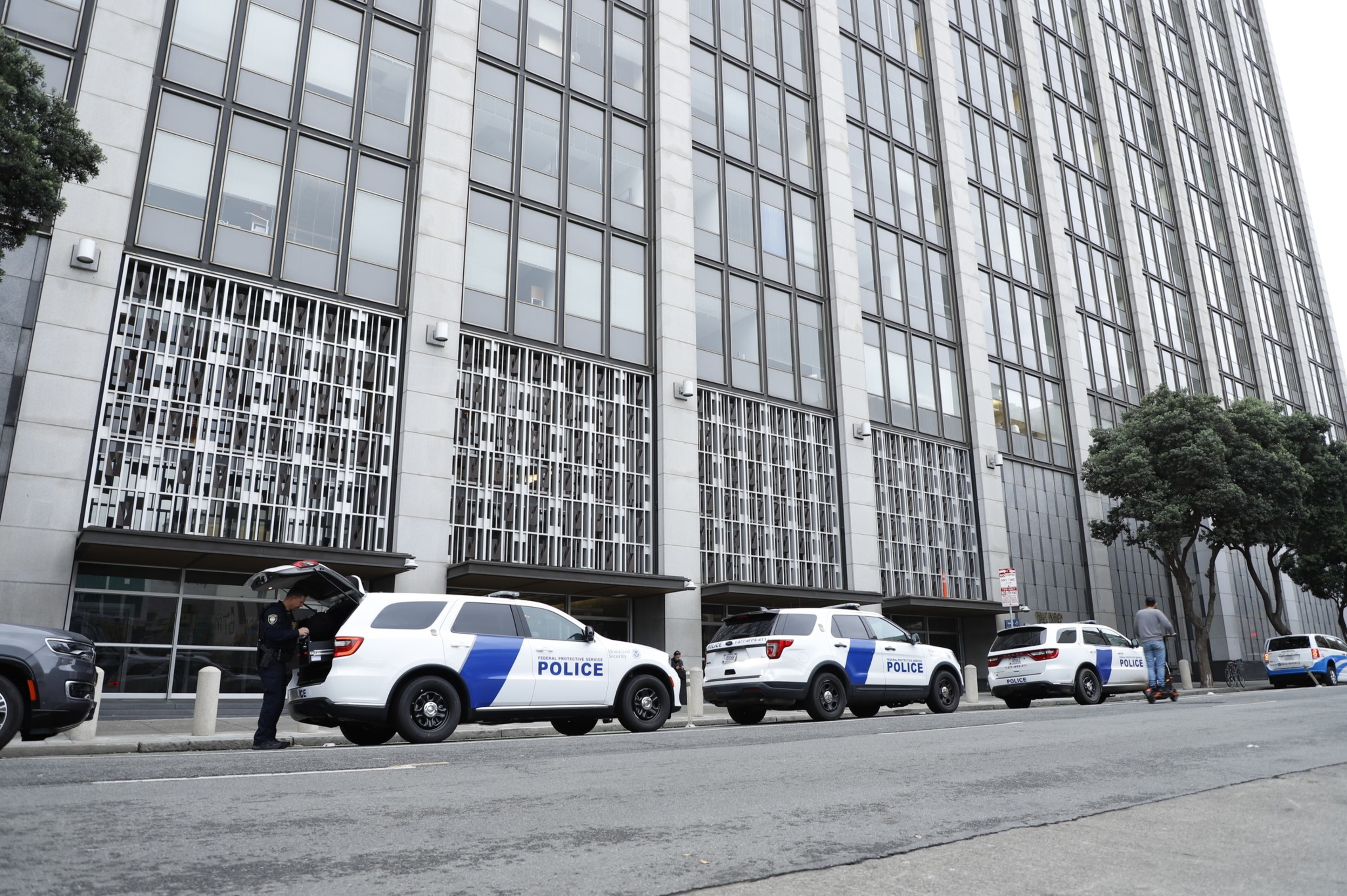 A row of police SUVs is parked in front of a large, multi-story building. A person stands at one of the vehicles with its rear hatch open, while another rides a scooter nearby.