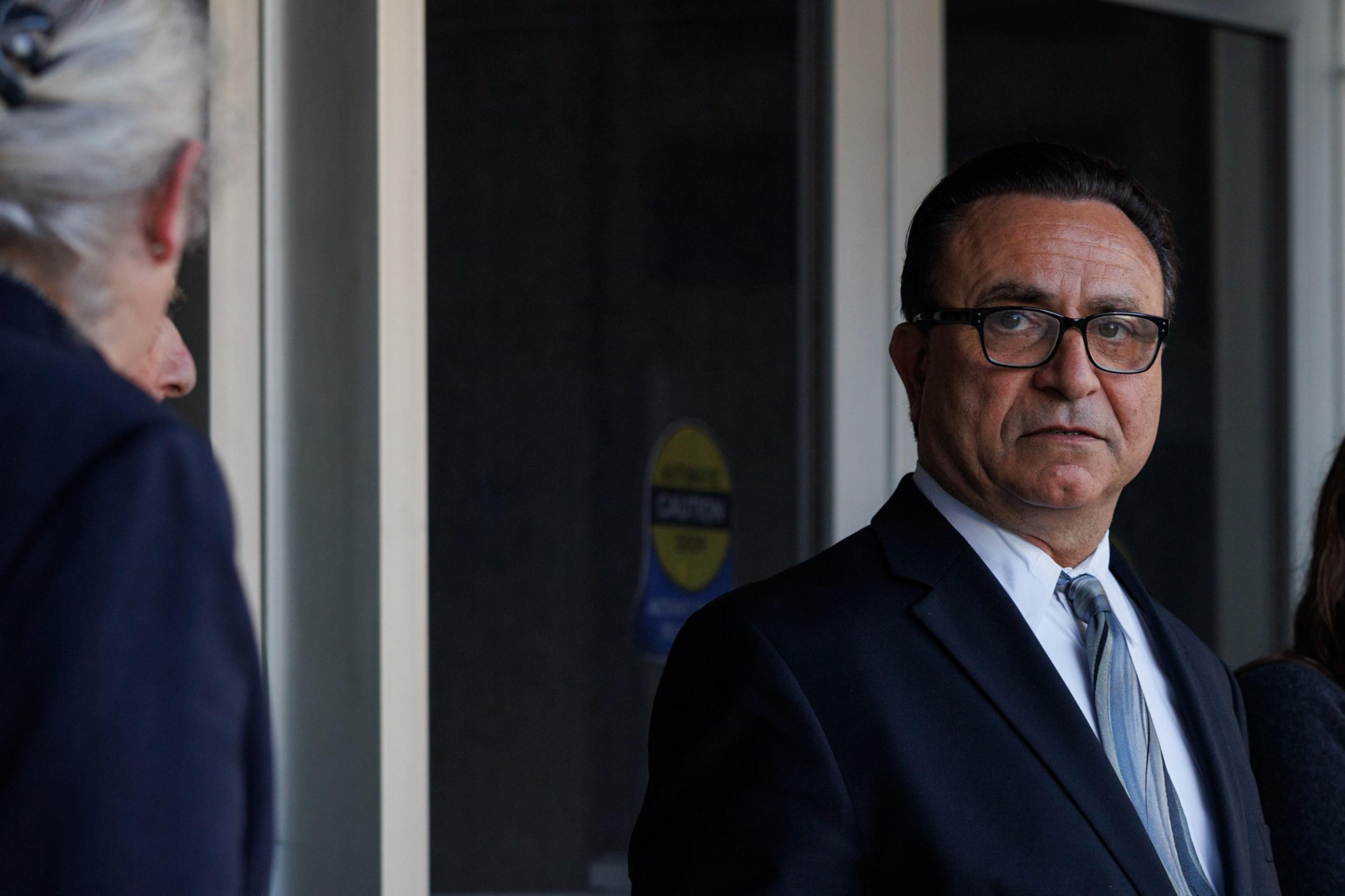 A man in a suit with glasses stands by a door, looking toward the camera, with a blurry person in foreground.