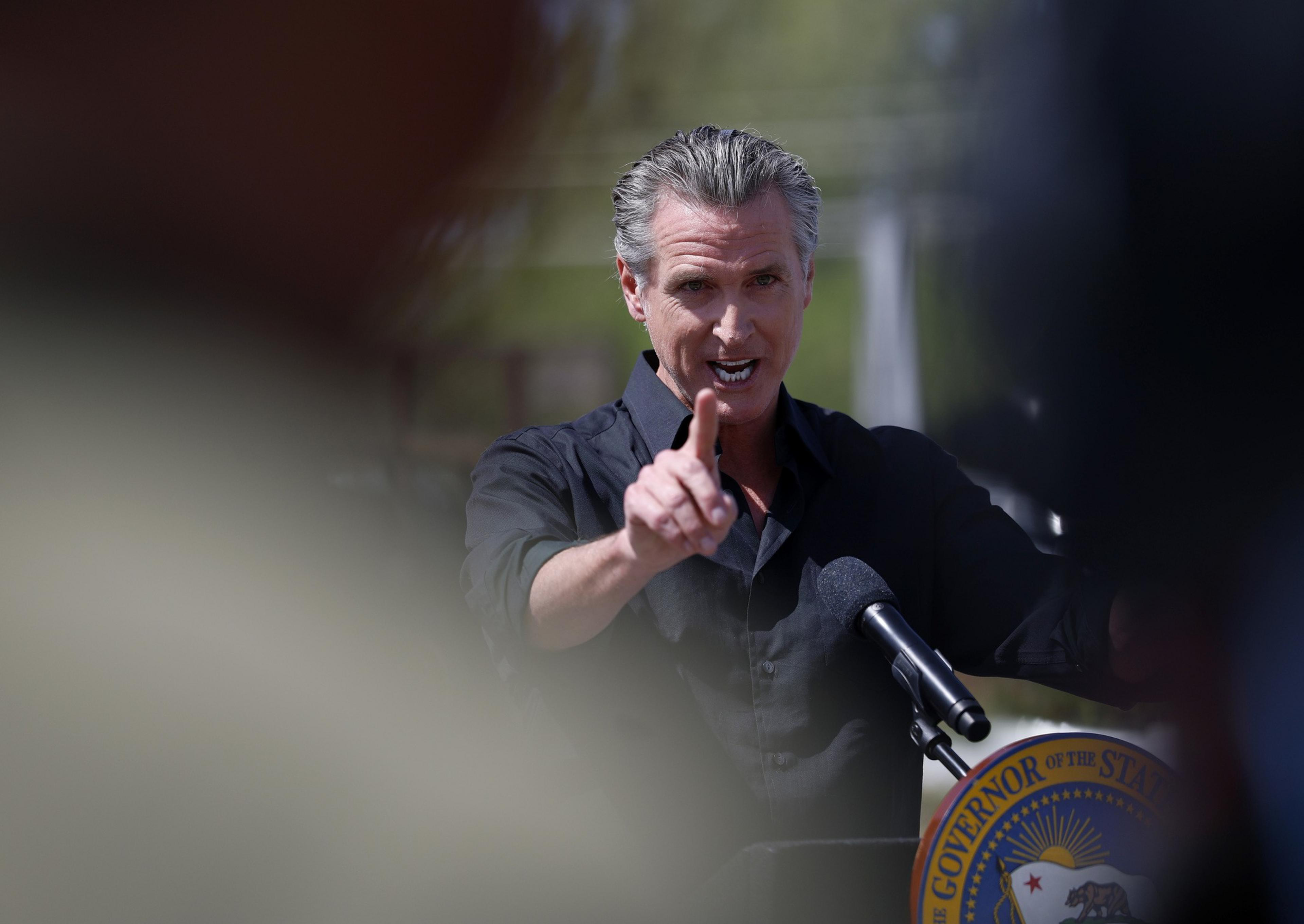 A person in a black shirt speaks passionately at a podium outdoors, pointing forward. A seal with "Governor of the State" is visible on the stand.