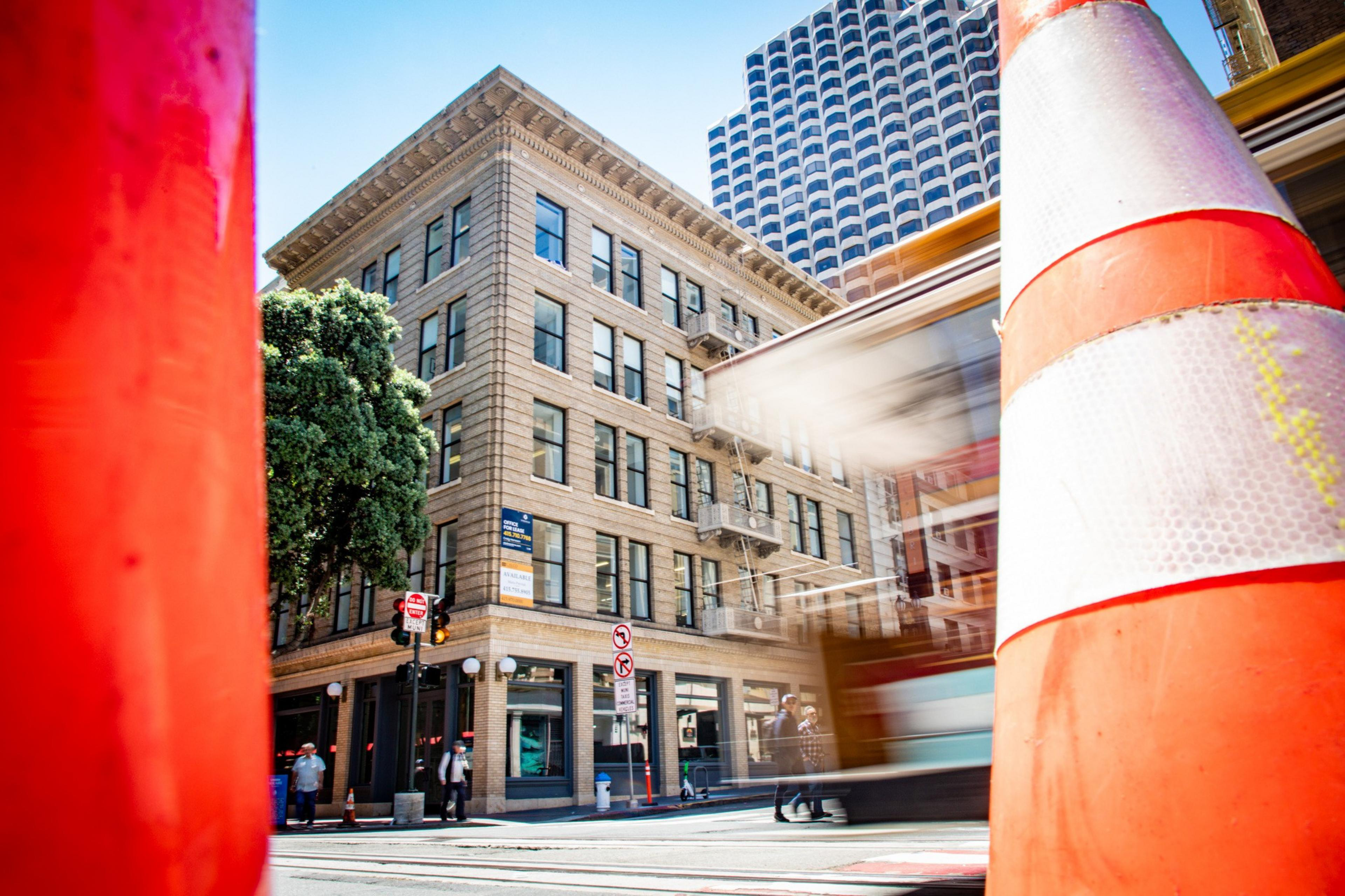 A brick corner building with large windows is seen between two blurry moving orange cones. A cable car passes by, and a modern skyscraper stands in the background.