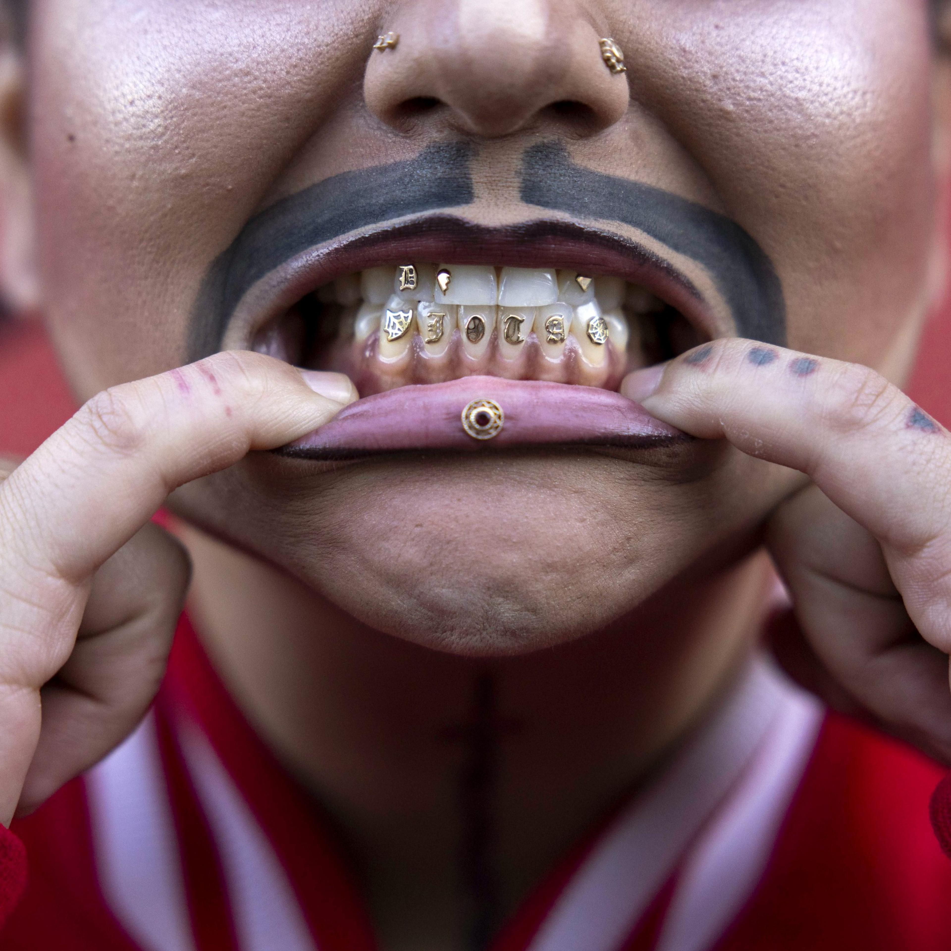 A person with tattoos and visible piercings is pulling down their lower lip to show gold-plated teeth and a gold piercing in the lower lip.