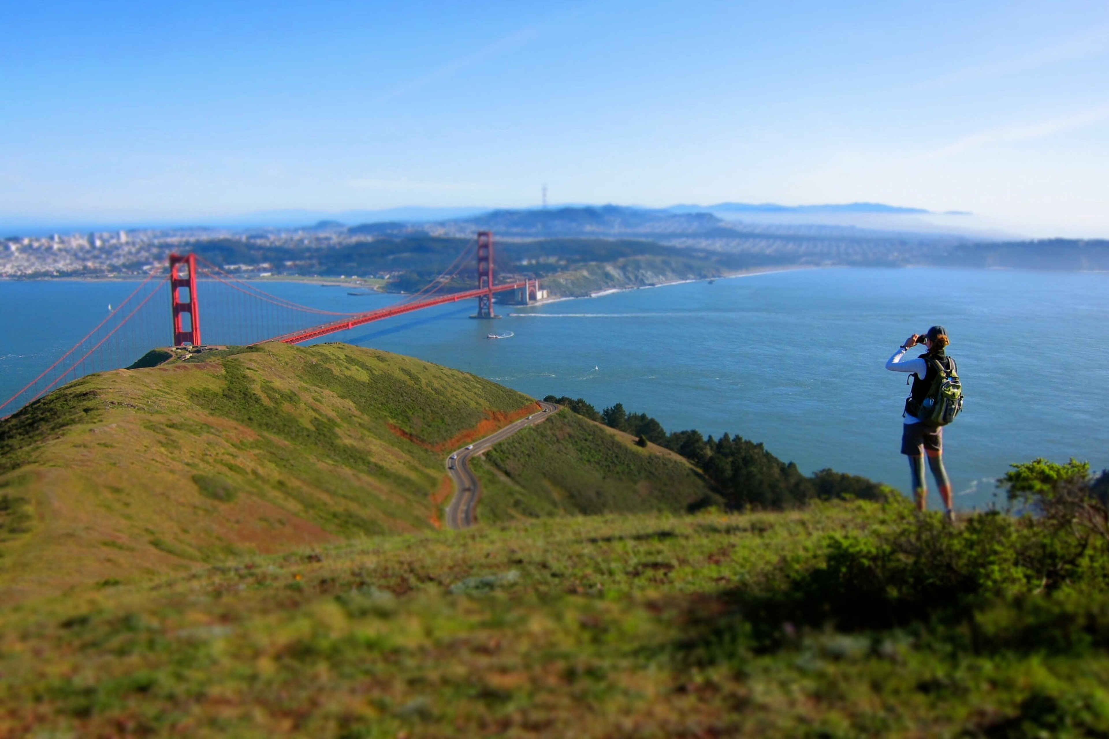 A person with a backpack stands on a grassy hill overlooking the Golden Gate Bridge stretching across a blue bay under a clear sky.