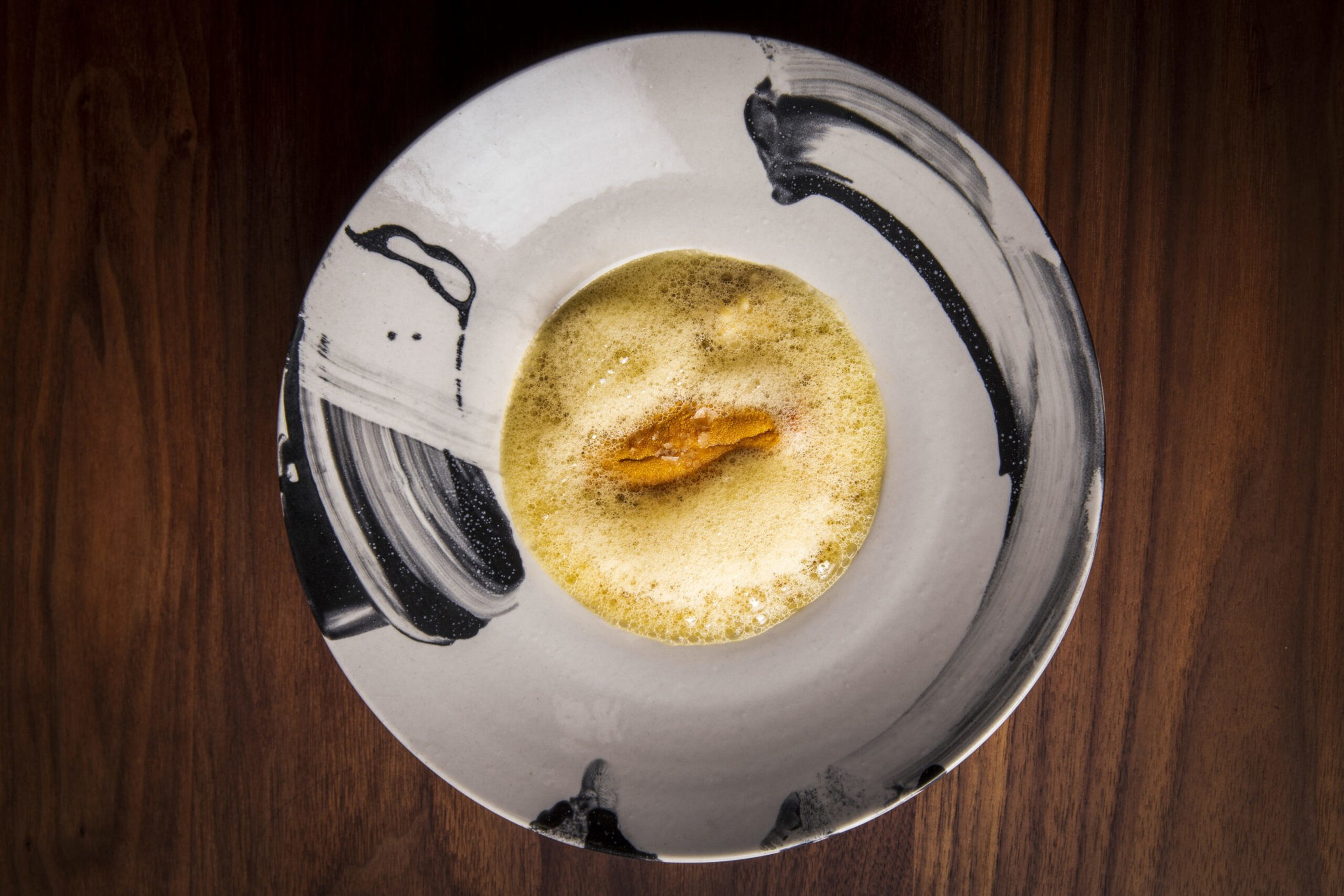 A white plate with black abstract brush strokes holds foam surrounding a small piece of golden-brown food in the center, set on a wooden surface.