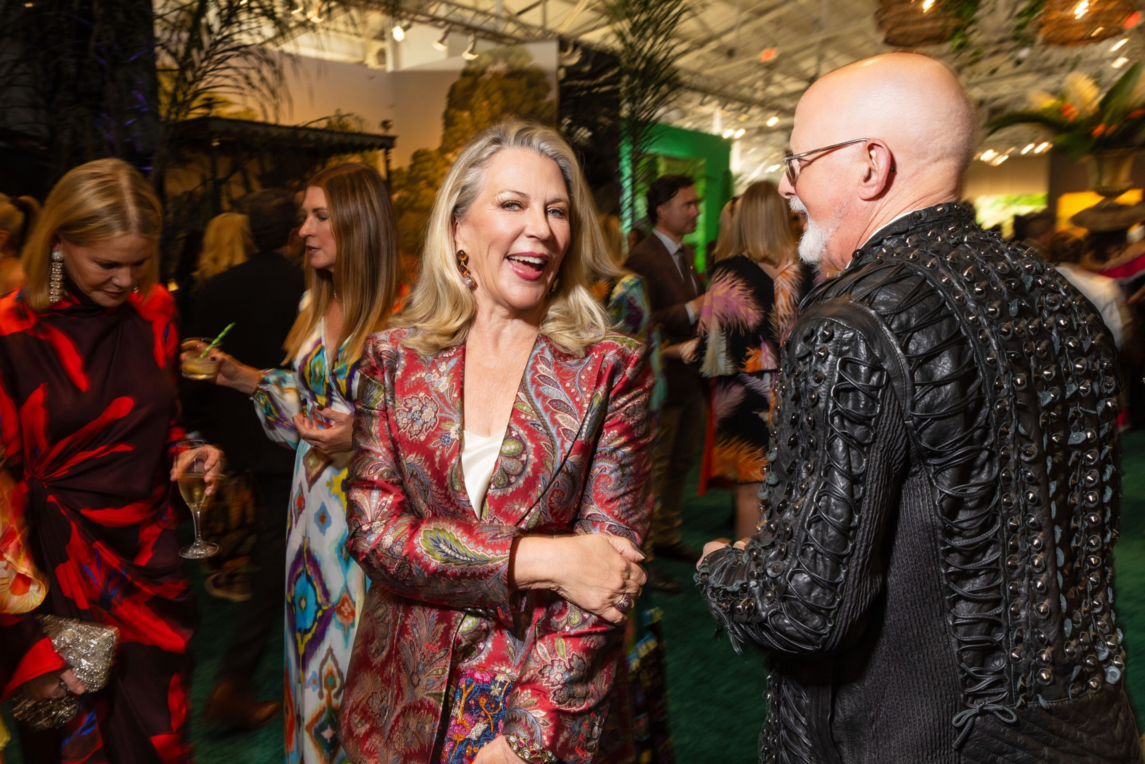 A woman in a red paisley jacket laughs while talking to a bald man in a black studded jacket at a lively indoor event with other guests.
