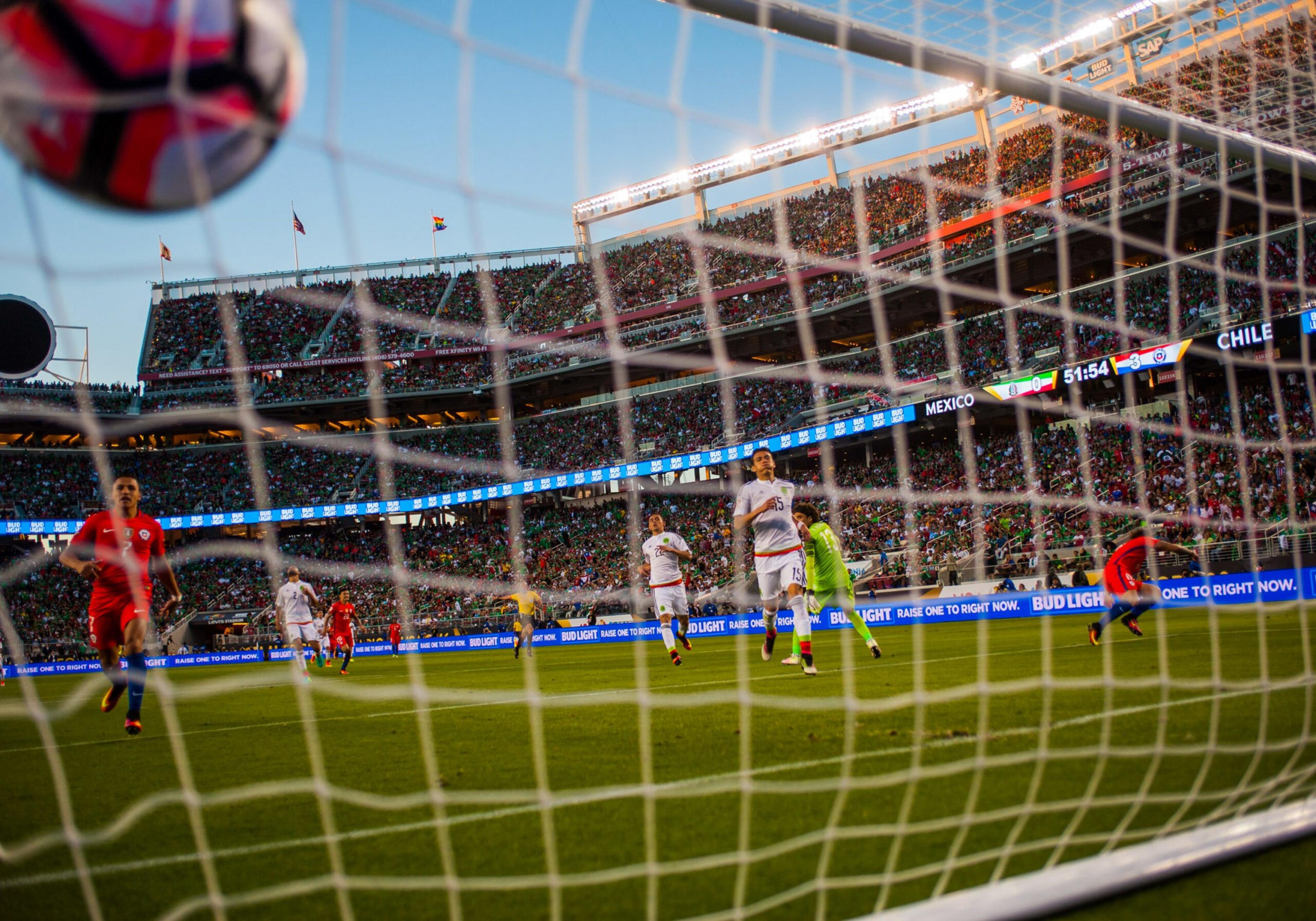 A soccer match in progress, viewed from behind the goal, with players on the field and a crowded stadium in the background.