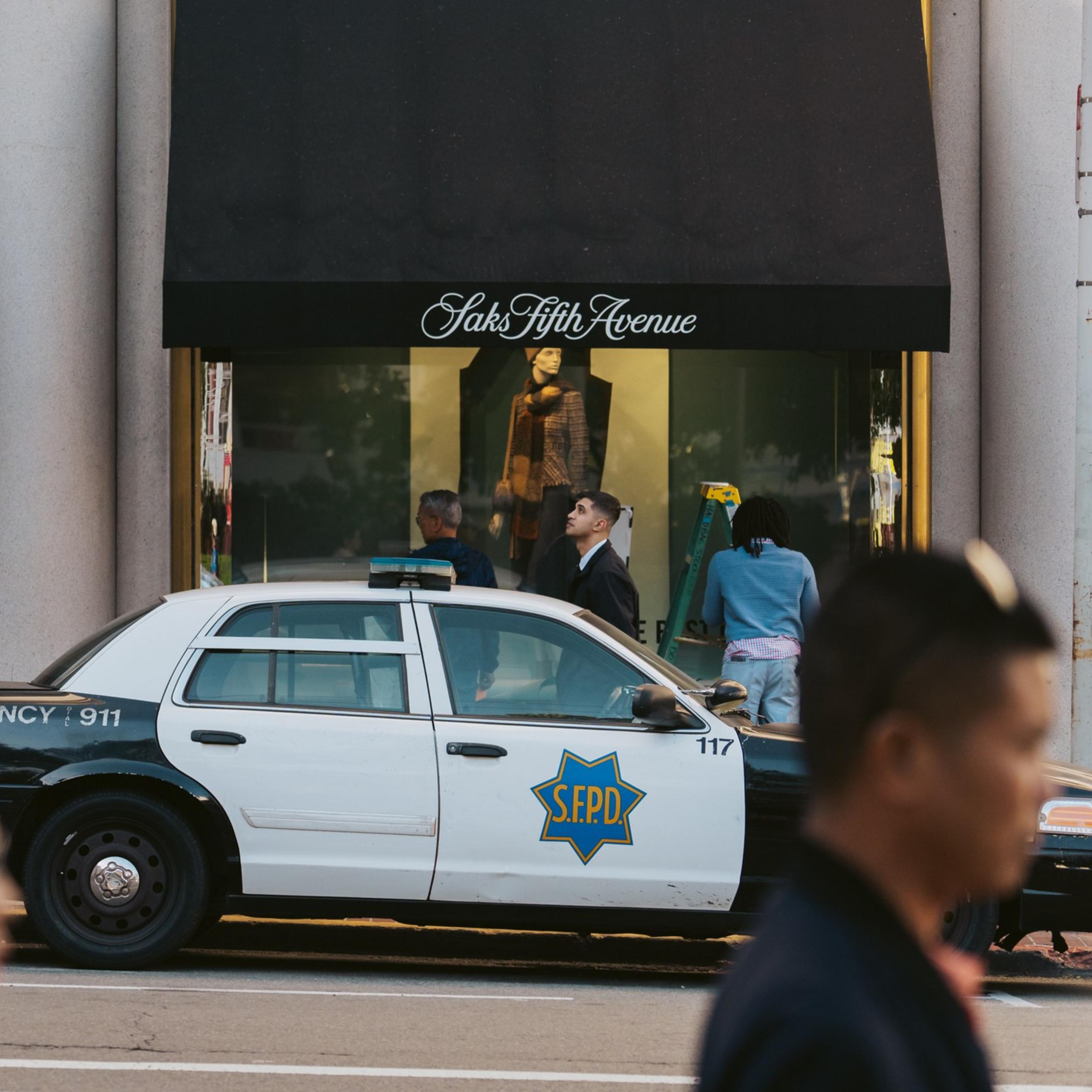 A police car is parked outside a Saks Fifth Avenue store with people visible in front of the windows and a blurred passerby in the foreground.