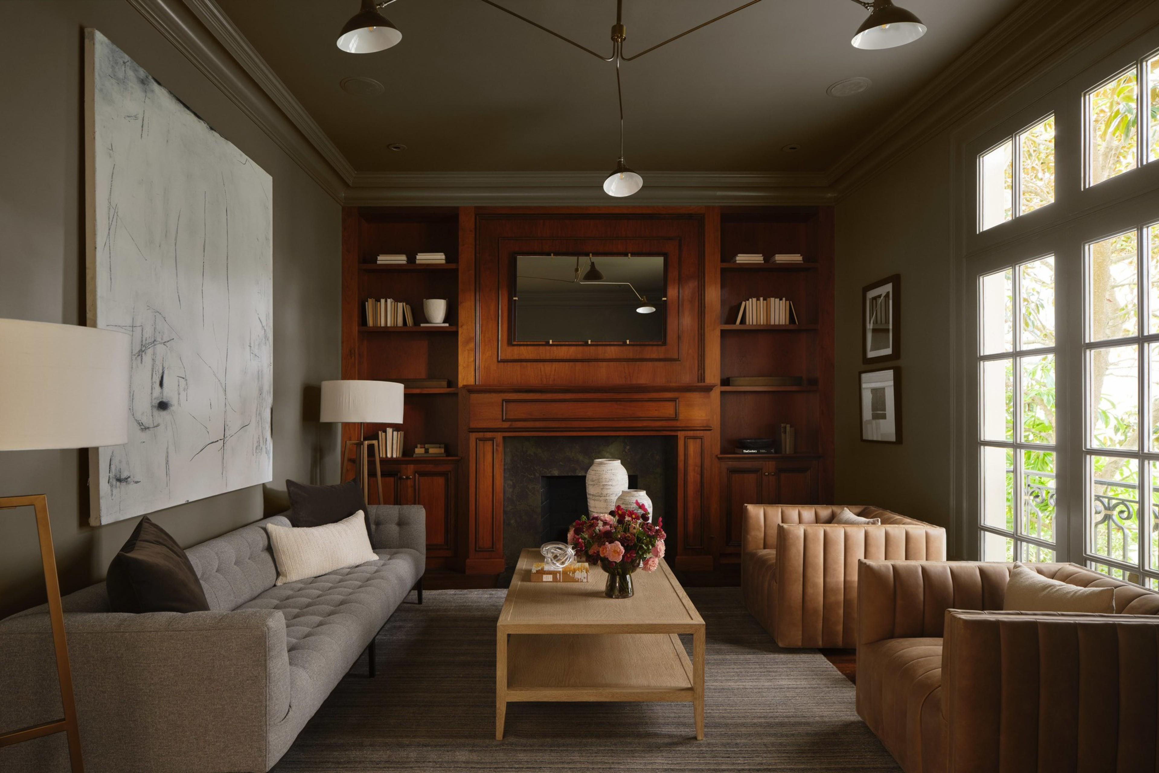 A cozy living room with a gray sofa, two tan armchairs, a wooden coffee table with flowers, a dark wood fireplace, and large windows letting in natural light.