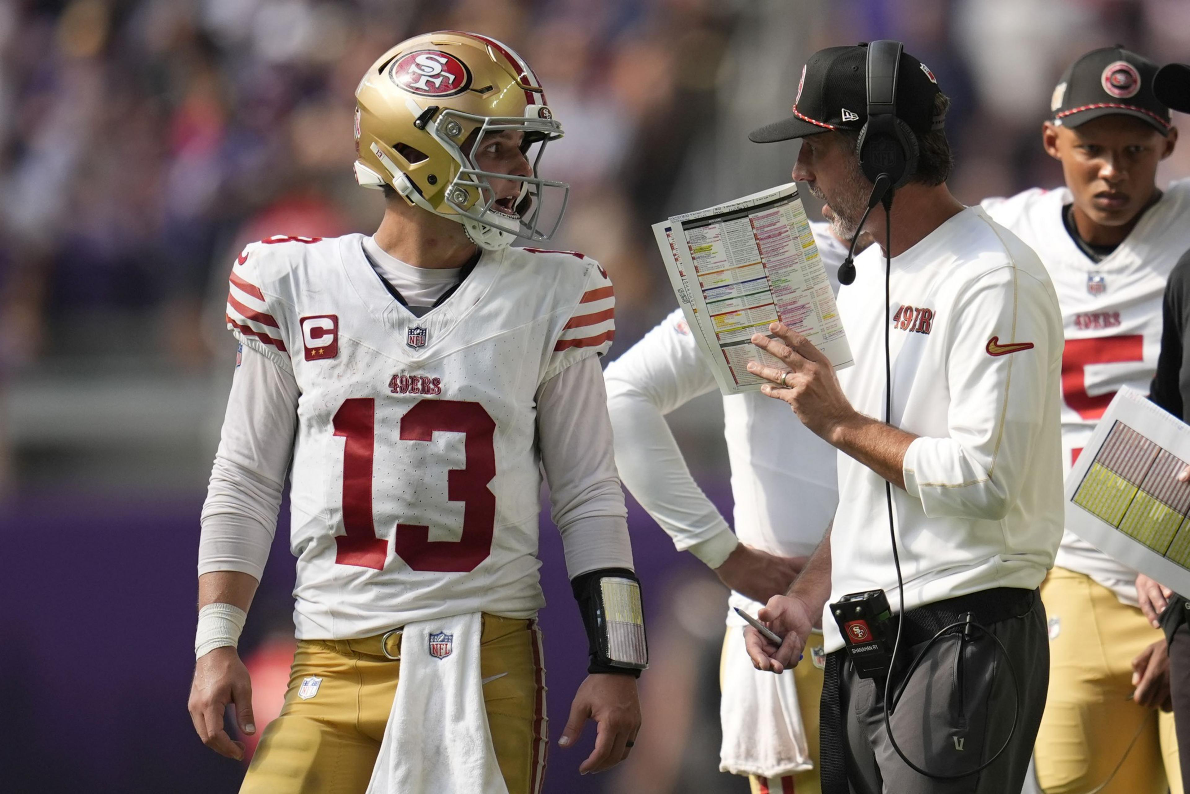 A football player in a white jersey and gold pants with a helmet listens to a coach holding a play sheet. Another person in uniform is in the background.