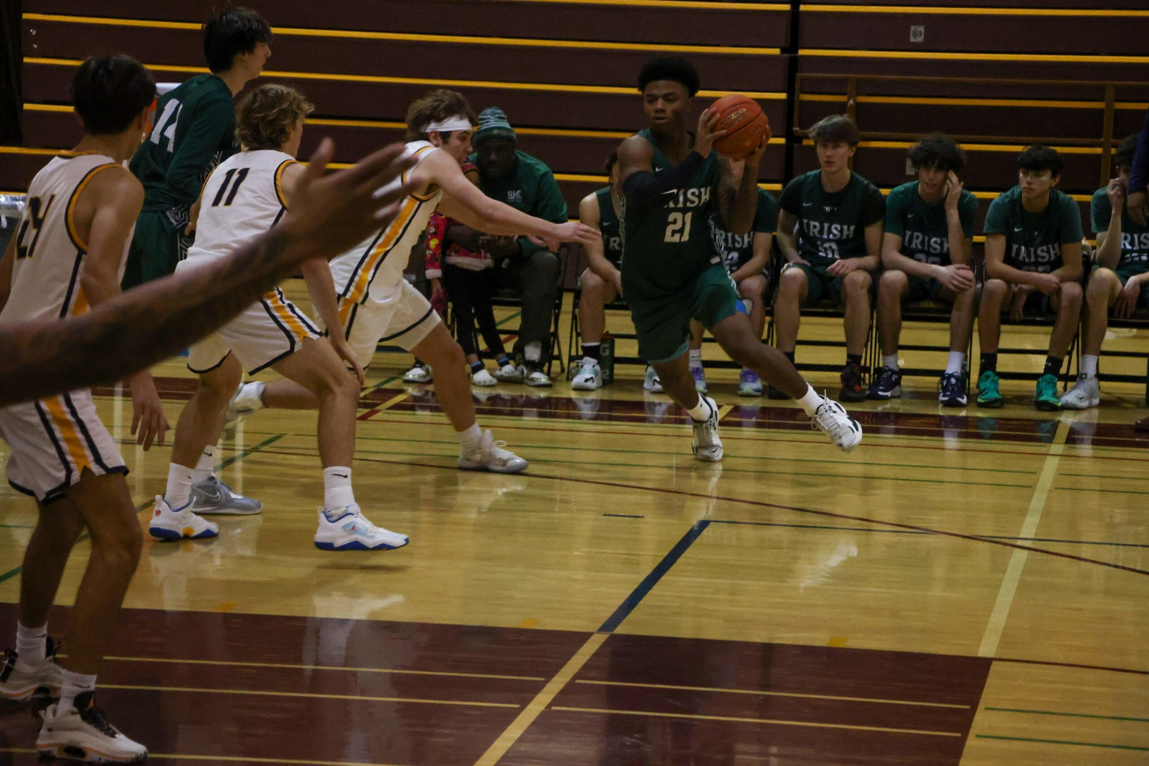 A basketball player in green, number 21, dribbles the ball while white team players rush to defend, with teammates and coaches seated in the background.