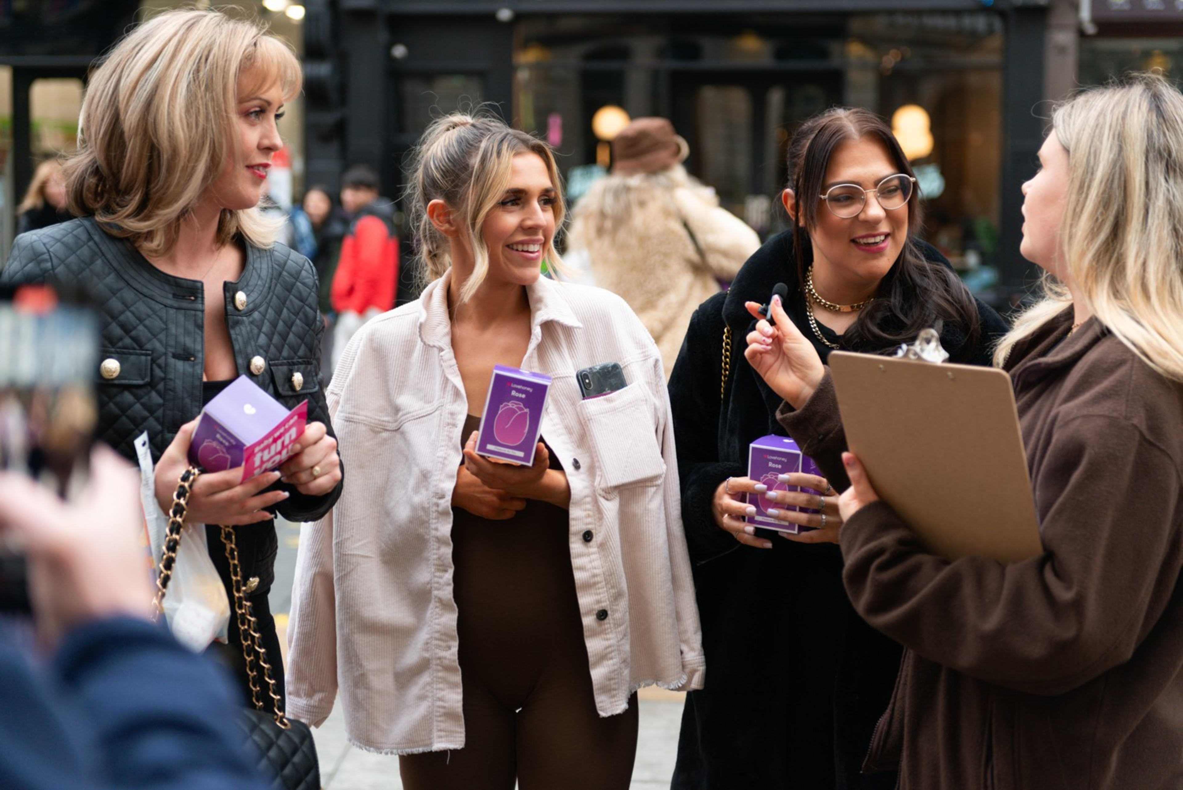 Four women are standing on a street, talking and holding purple boxes. One holds a clipboard and appears to be leading a conversation.