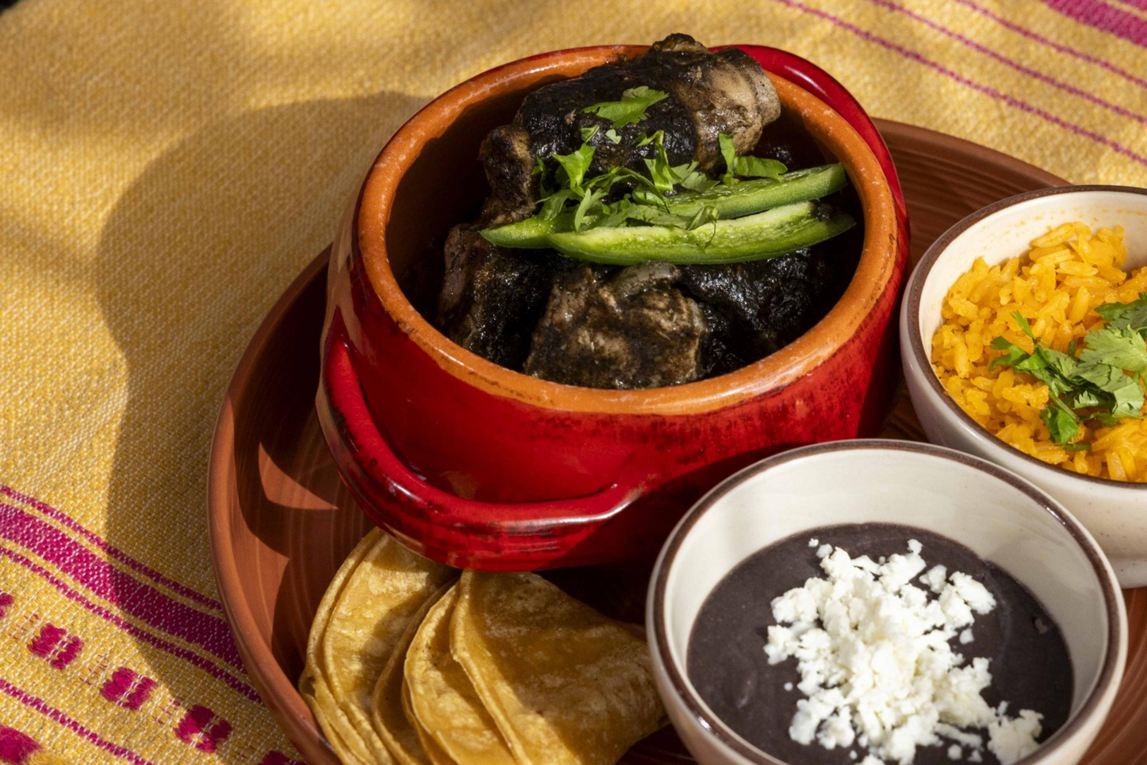 A plate features a red pot of dark stew with herbs, two bowls with yellow rice and black beans topped with cheese, and folded tortillas on a colorful cloth.