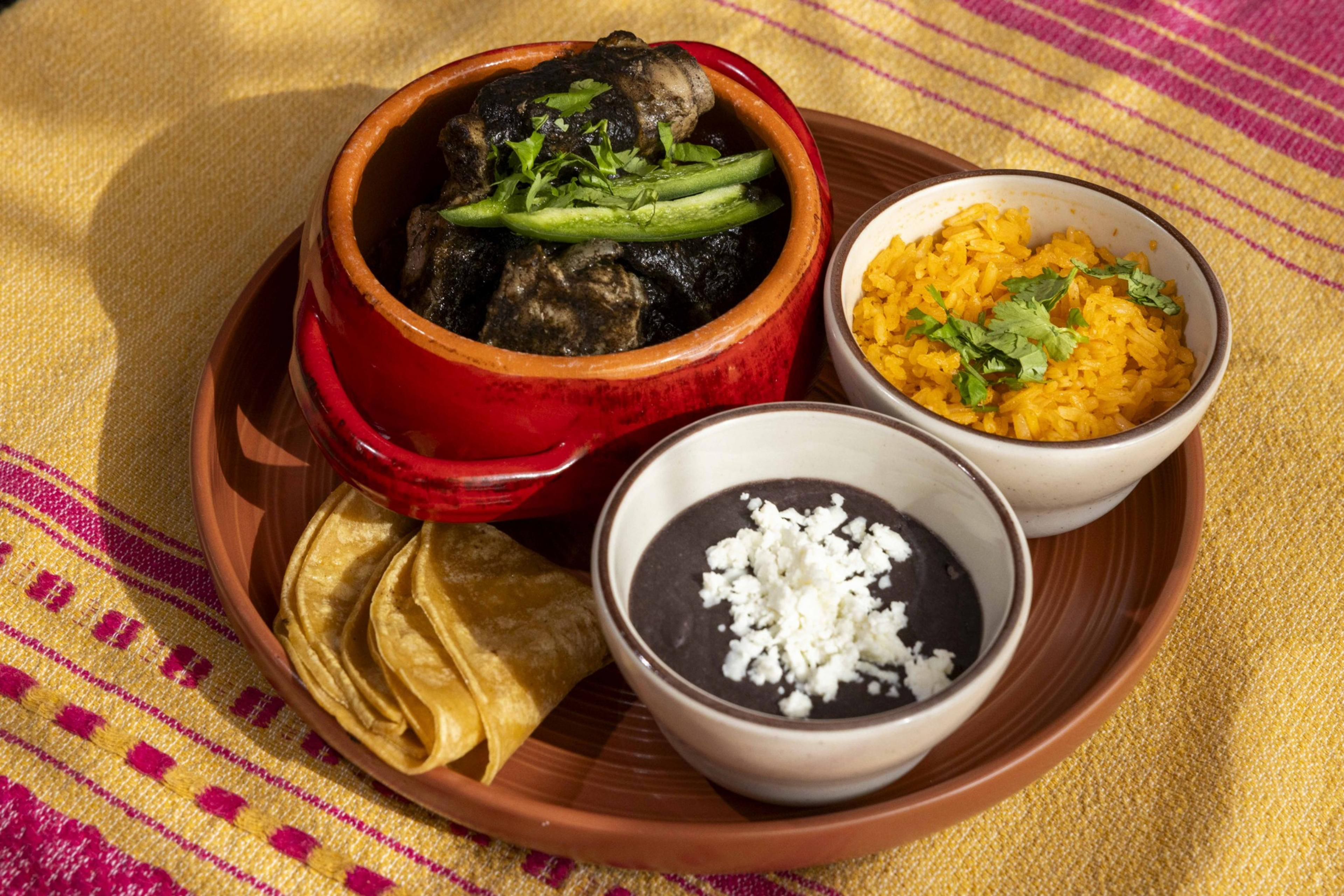 A plate features a red pot of dark stew with herbs, two bowls with yellow rice and black beans topped with cheese, and folded tortillas on a colorful cloth.
