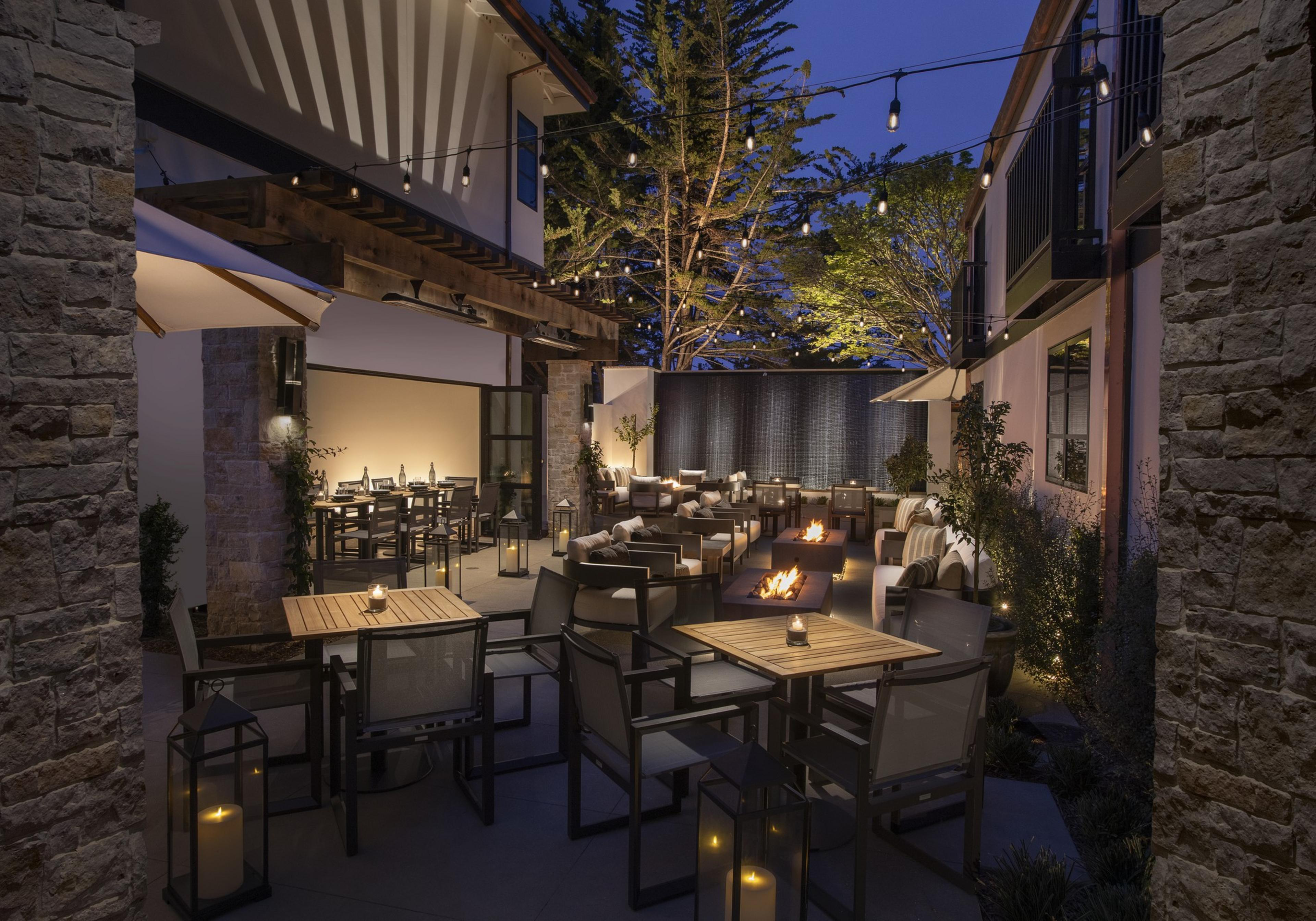 An enchanting outdoor patio scene at night is illuminated by string lights, with cozy seating, tables set with candles, and warm fire pits. Trees frame the background.