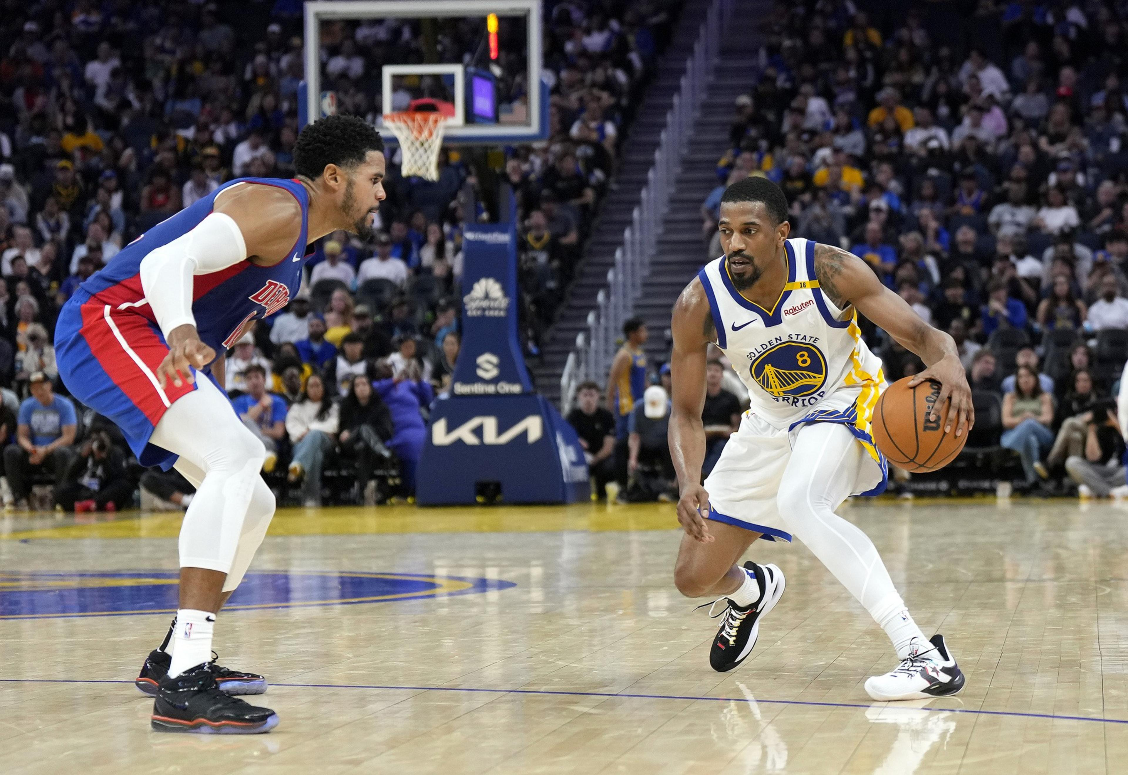 A basketball player in a Golden State Warriors uniform dribbles the ball while being guarded by a player in a red and blue uniform on a well-lit court.