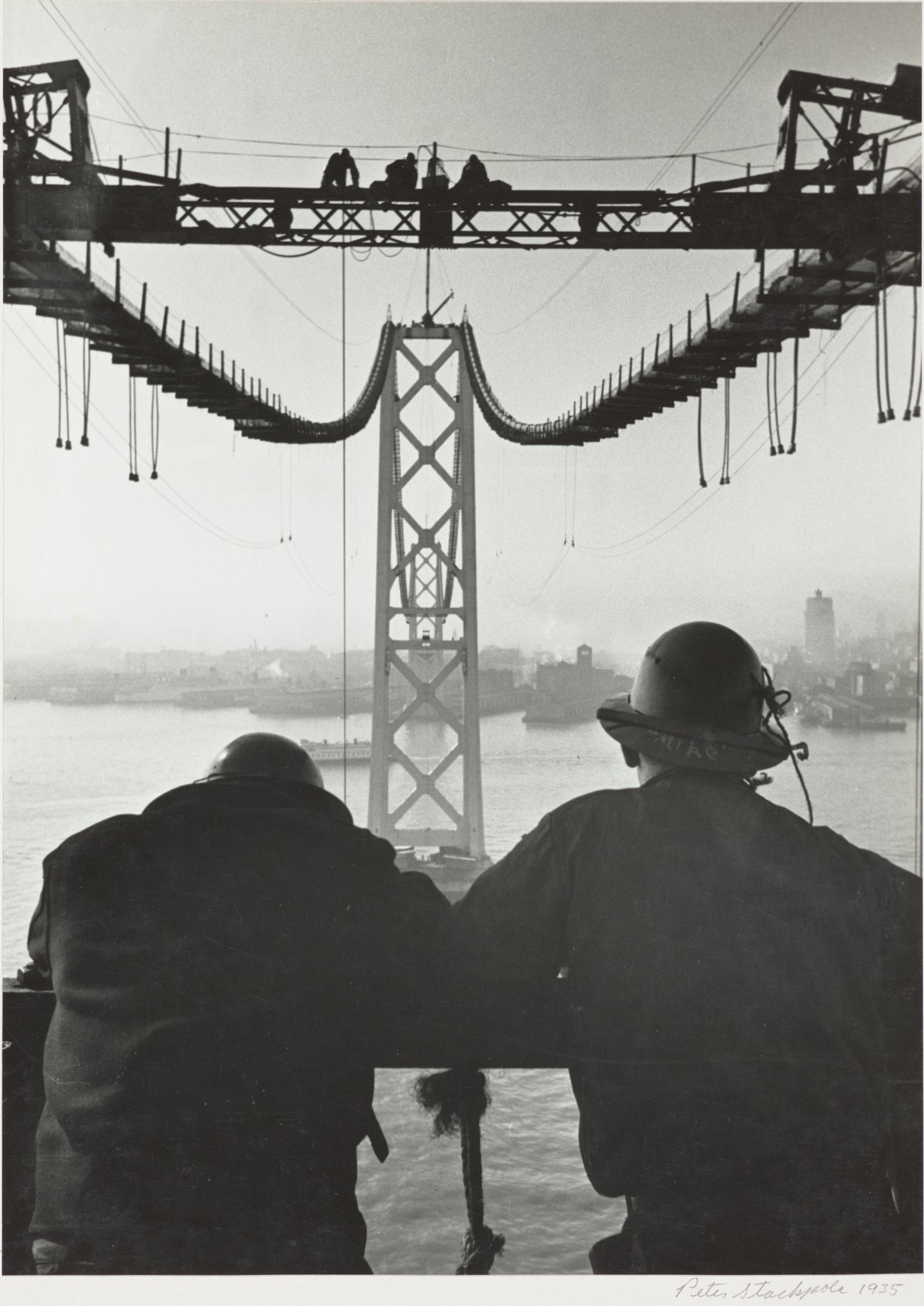 Two workers in helmets lean on a railing, overlooking the construction of a suspension bridge with a steel tower and cables above water.