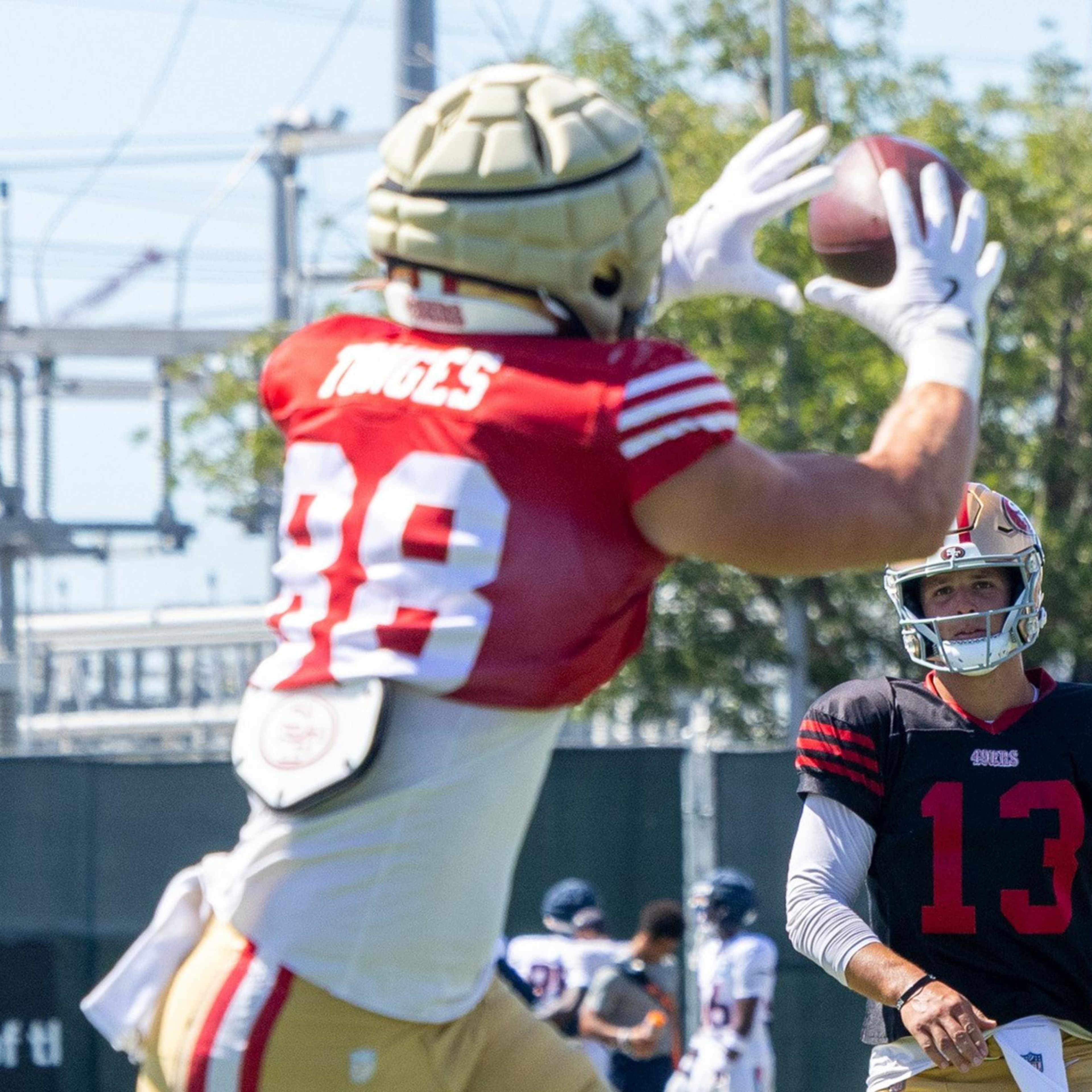 A football player in a red jersey and gold helmet catches a ball while another player in a black jersey and gold helmet watches during practice.
